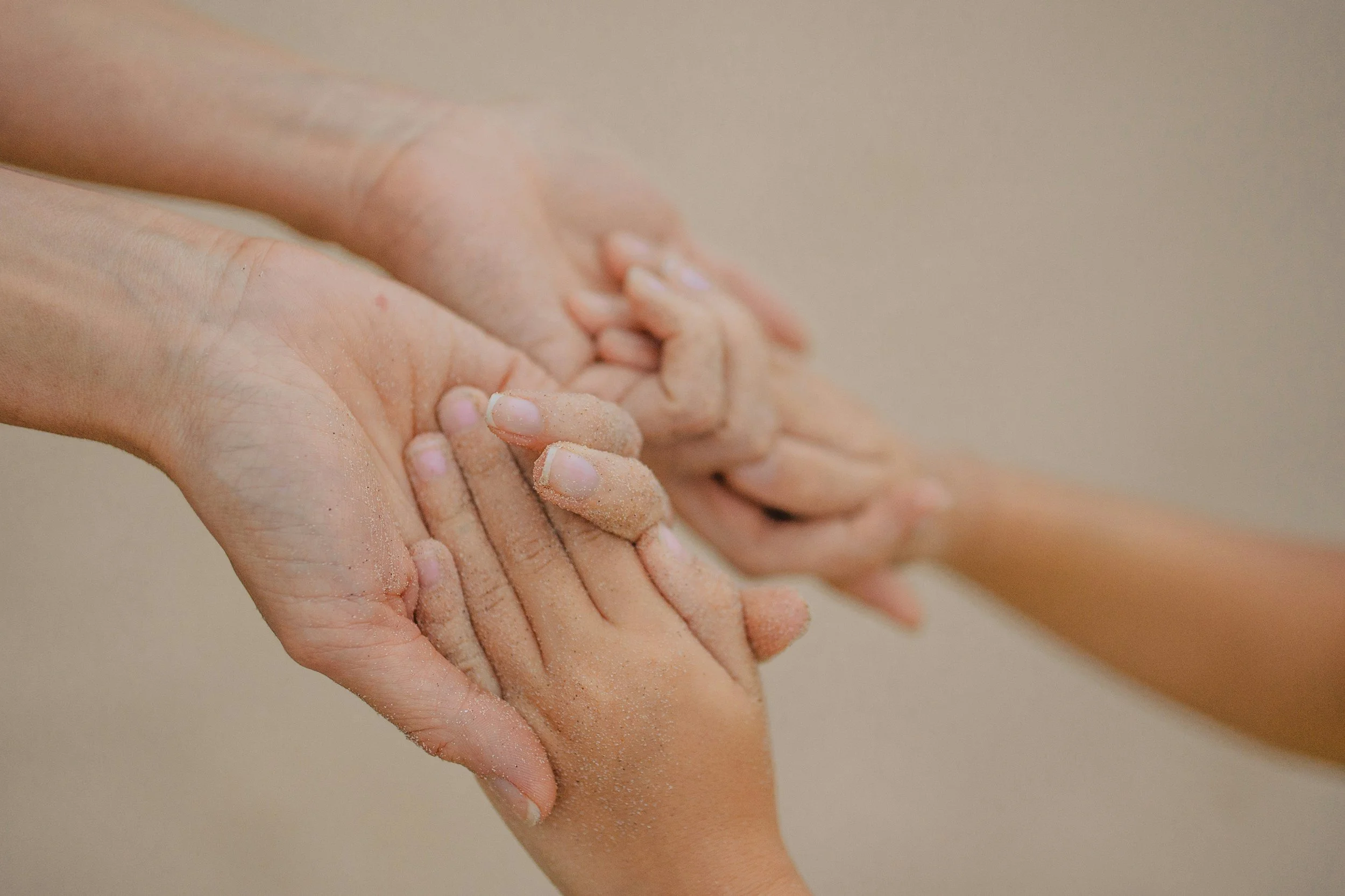 Due mani che si tengono, una più grande e una più piccola, entrambe coperte di sabbia, simboleggiando cura e protezione.