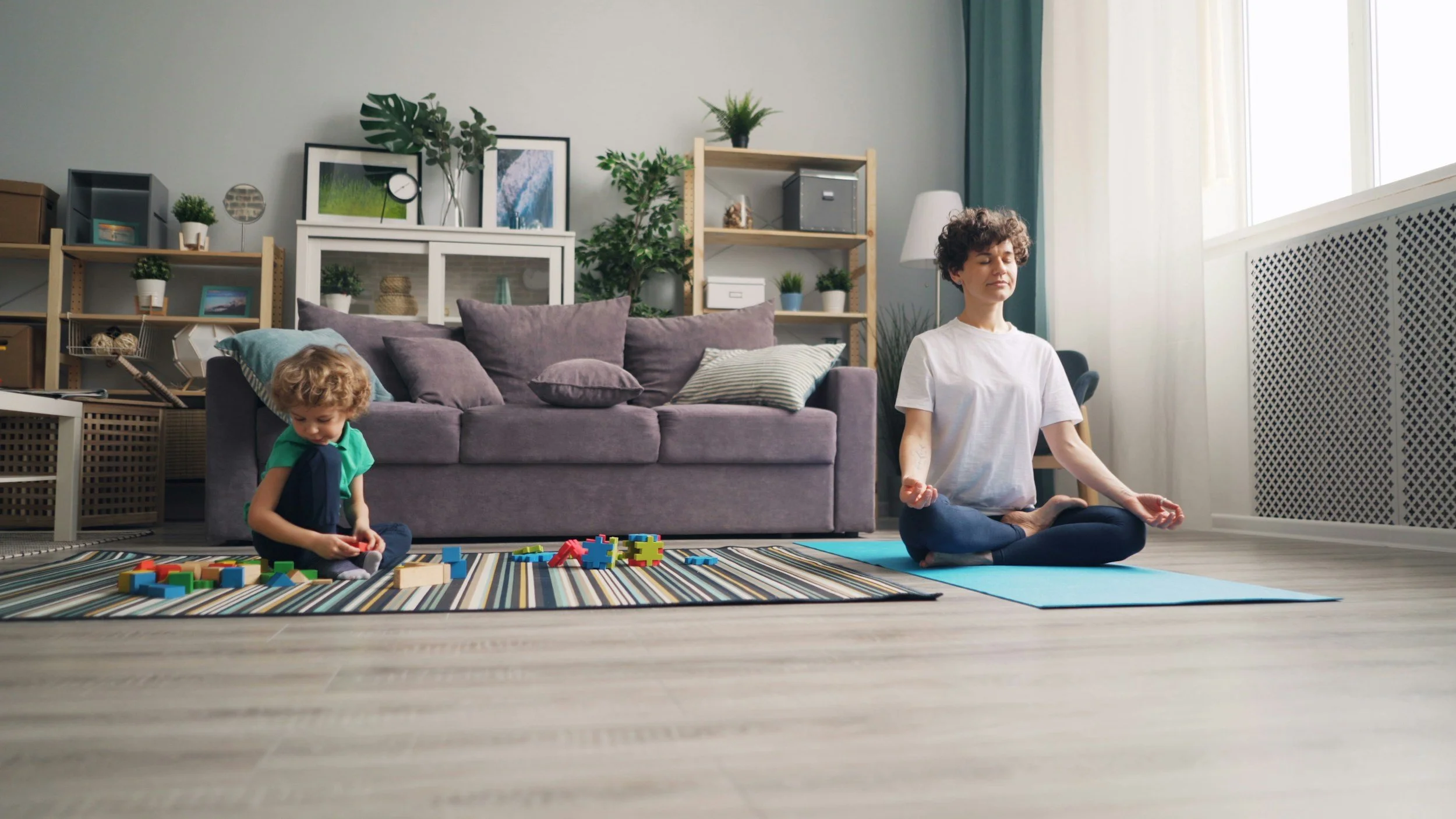 Una donna e un bambino asiatico fanno yoga e giocano con blocchi colorati su un tappeto nel soggiorno di casa, con scaffali pieni di piante e quadri sullo sfondo.