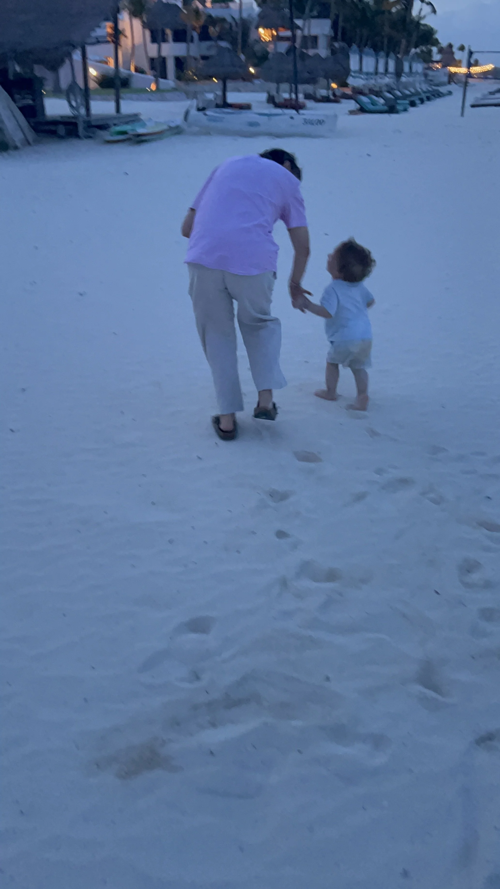 Un adulto e un bambino camminano insieme sulla sabbia bianca di una spiaggia al tramonto, con alcune barche ormeggiate sullo sfondo.