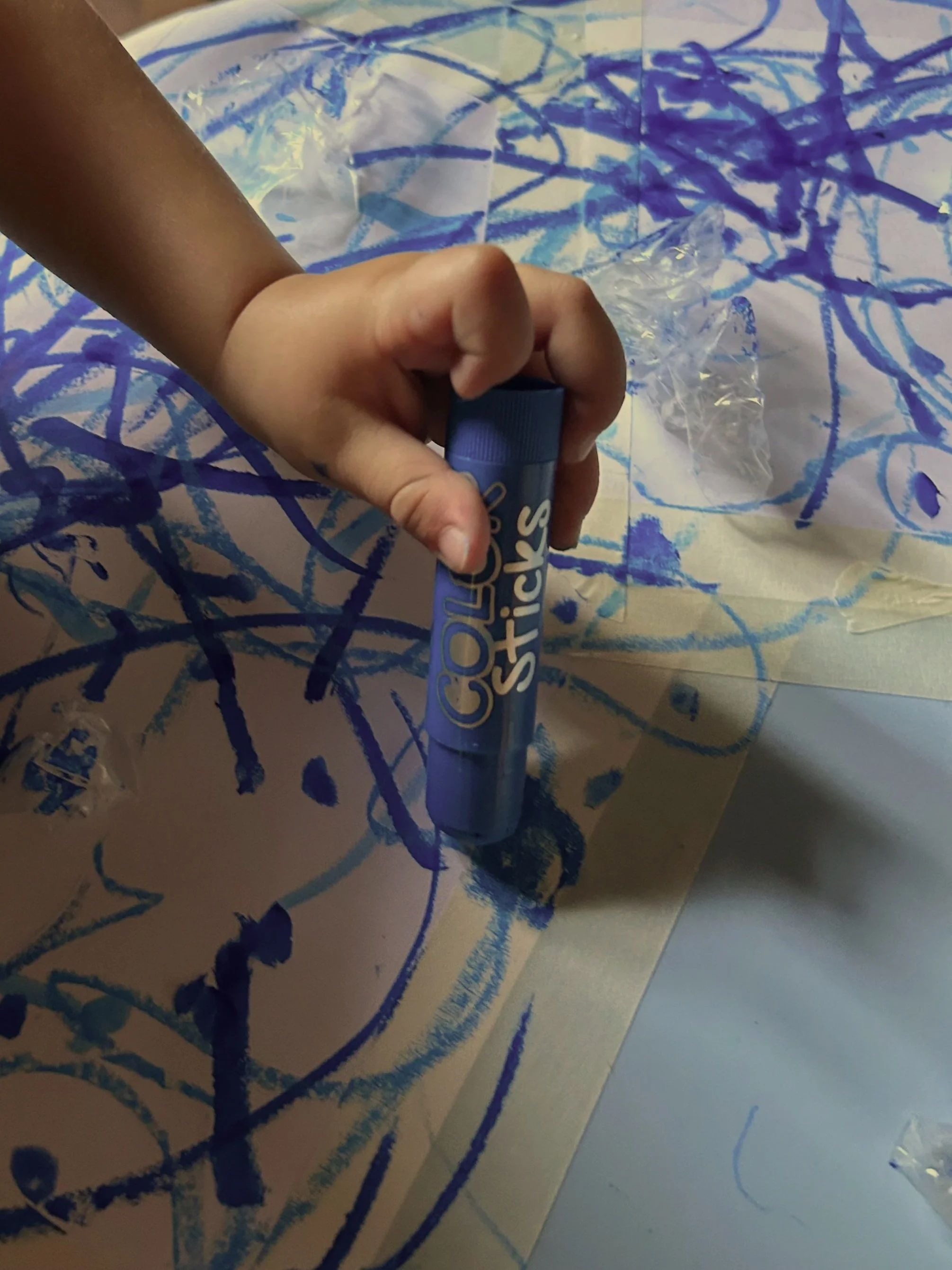 Mano che tiene un pastello blu mentre disegna su un foglio con linee blu e cristalli di plastica sulla superficie.