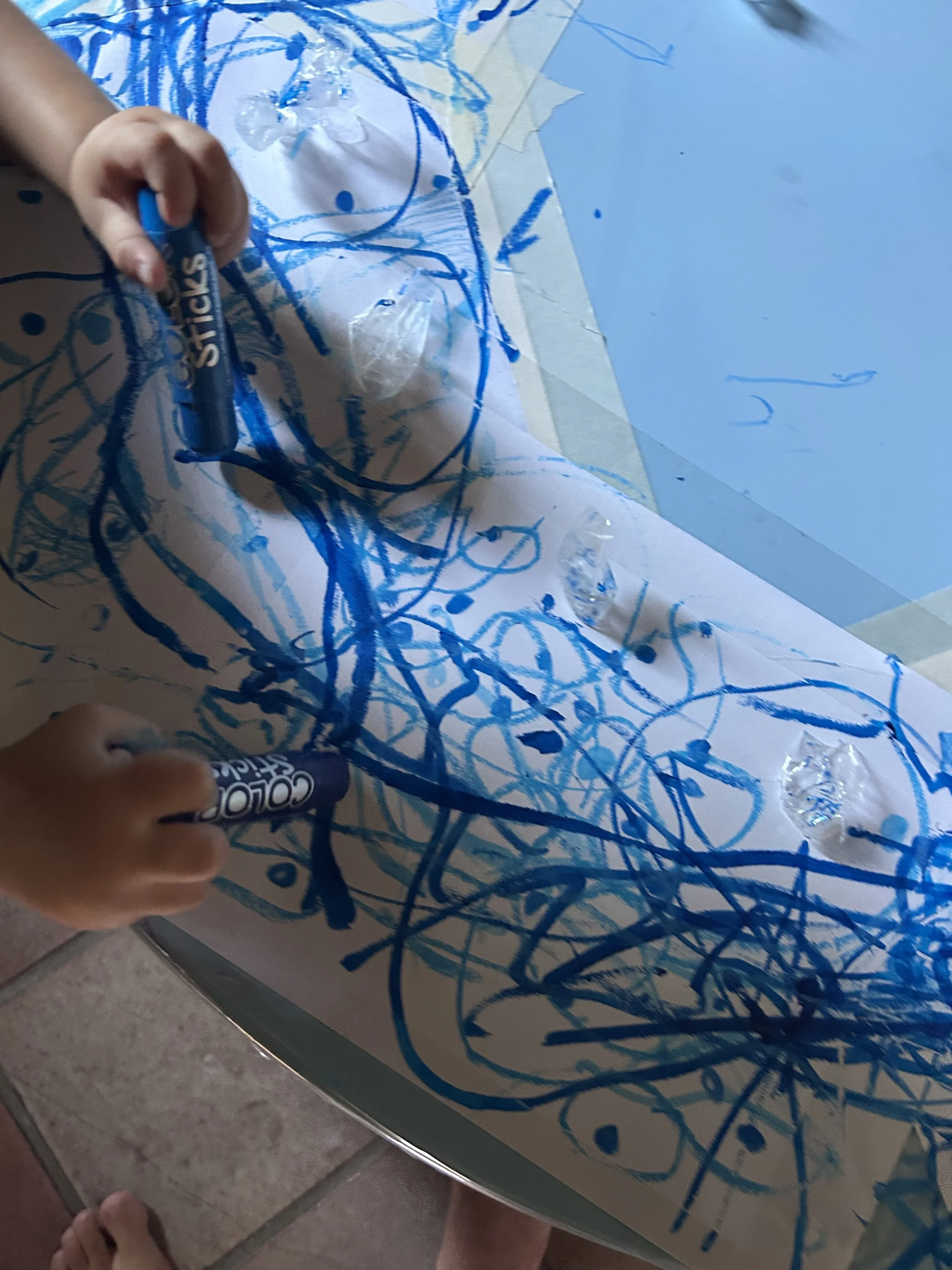 Bambino che disegna con un pennarello blu su una superficie di plastica trasparente, creando linee e forme astratte di colore blu.