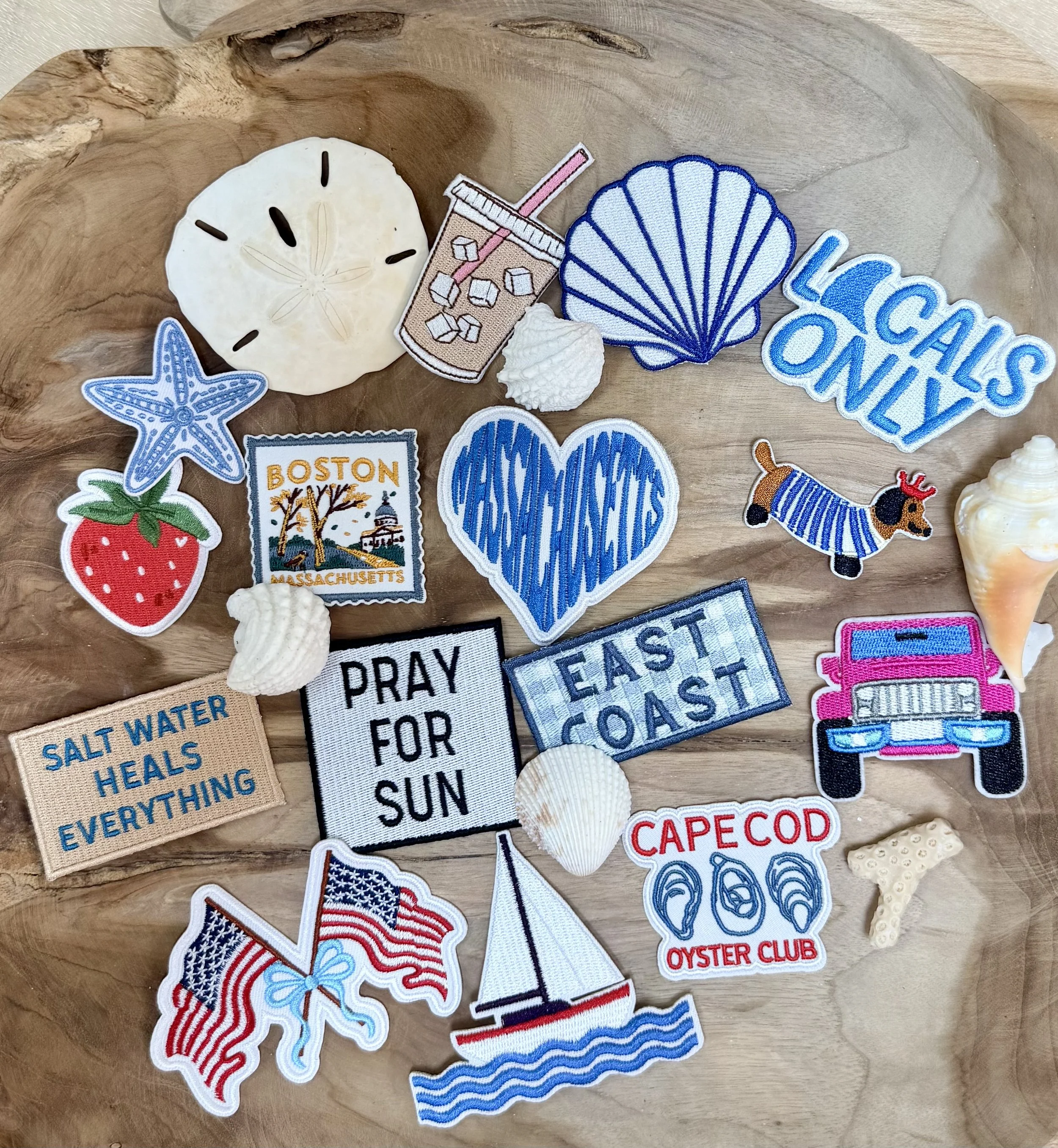 Collection of embroidered patches on a wooden surface with themes of summer, beach, and New England, including a starfish, strawberry, Boston Massachusetts, scallop shell, lobster, Cape Cod, oyster club, American flag, and a sailboat.