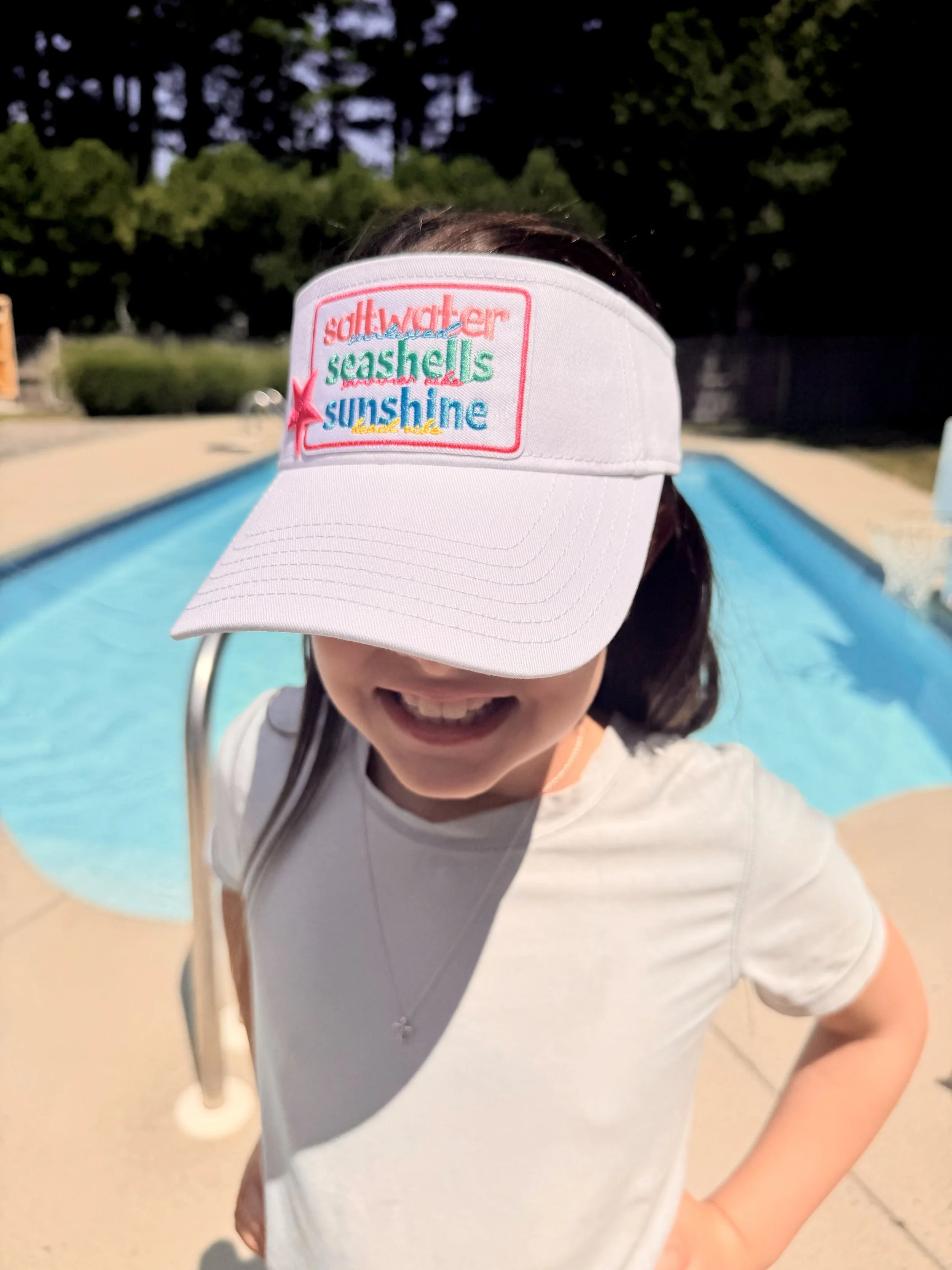 A person wearing a white cap with colorful embroidery, standing outdoors near a swimming pool.