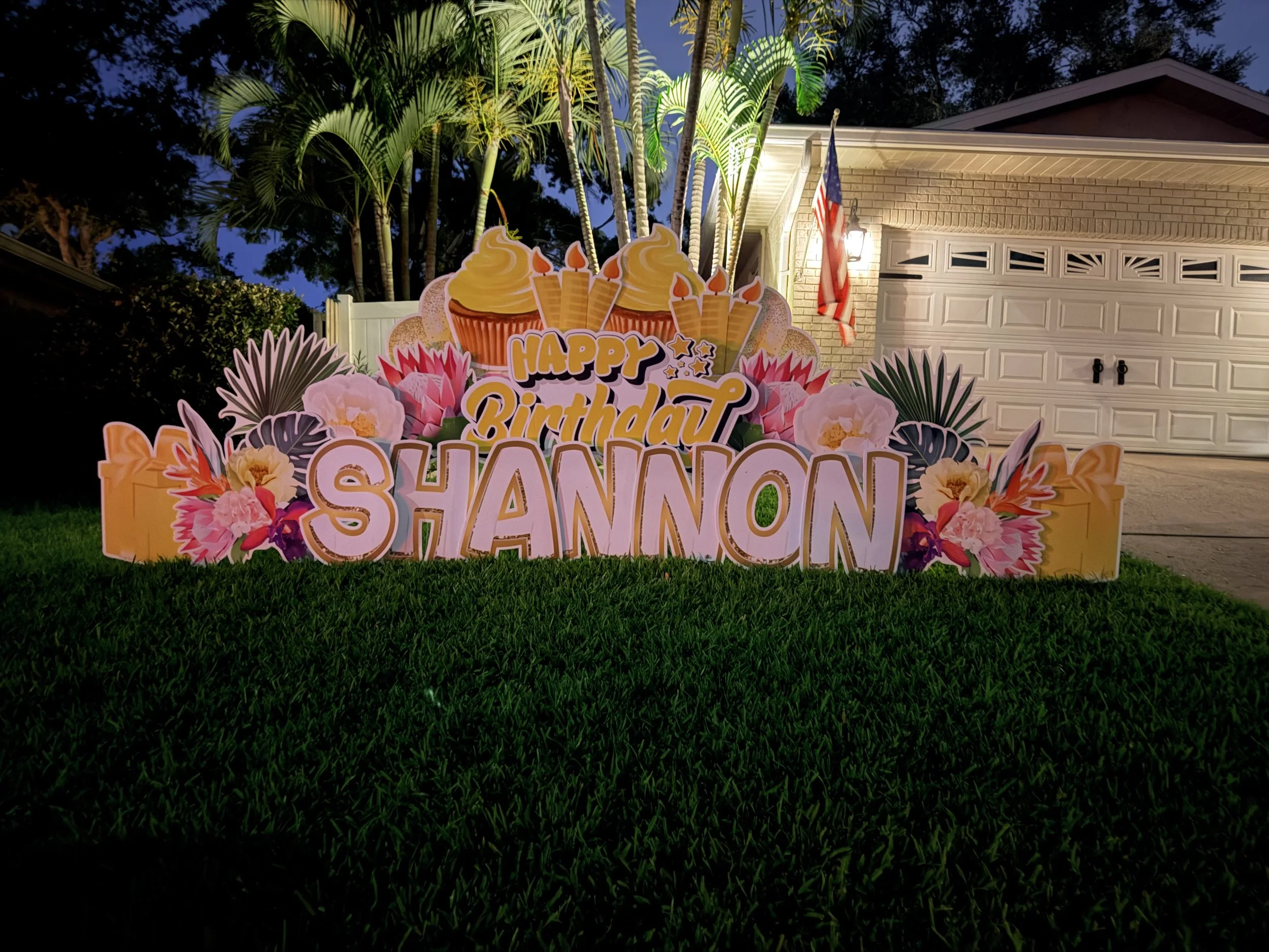 A decorated birthday sign with flowers, cupcakes, and candles, reading 'Happy Birthday Shannon,' in front of a house with garage and an American flag outside during evening.