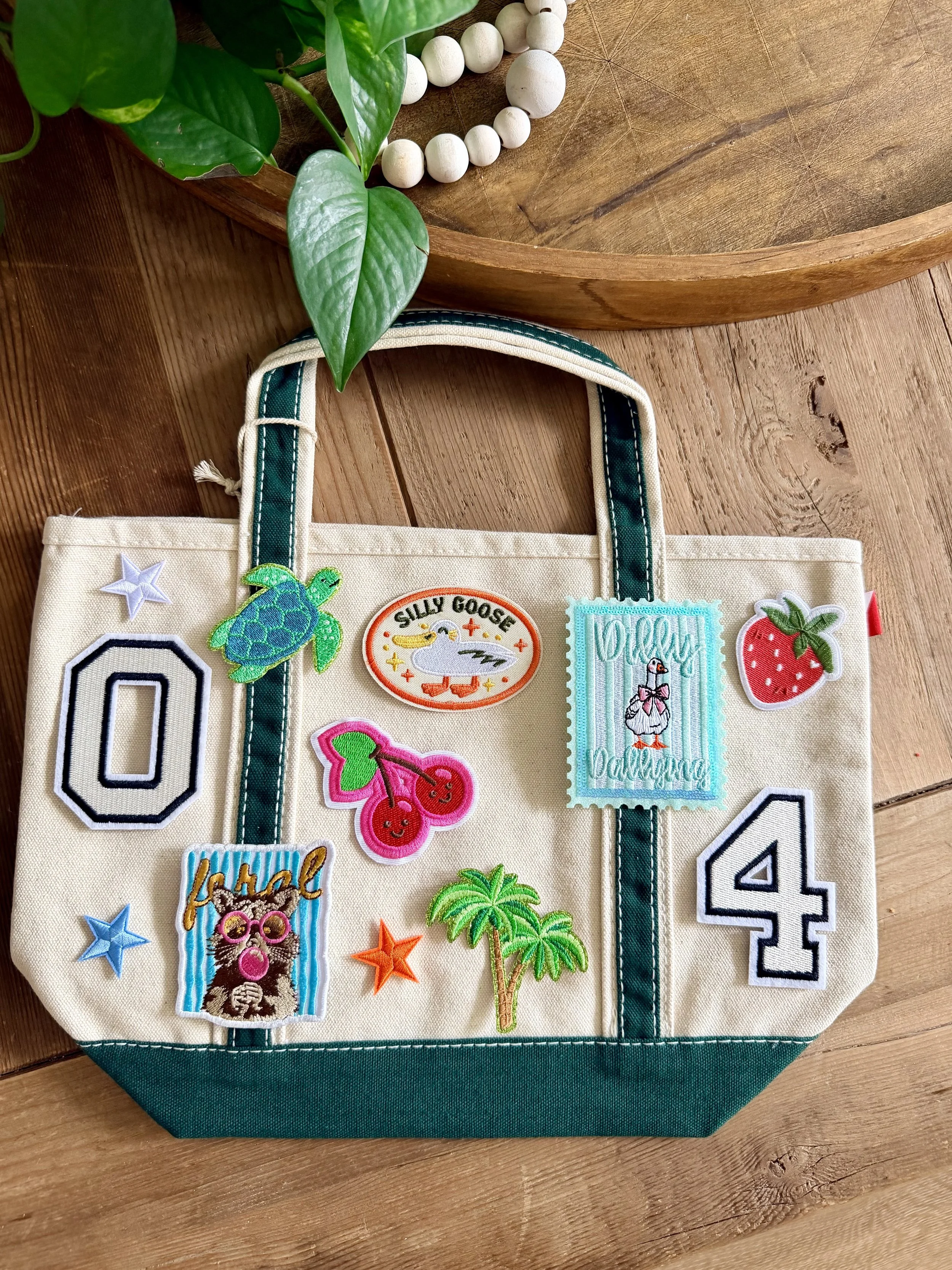 A fabric tote bag decorated with various colorful embroidered patches including a turtle, a duck, strawberries, a pineapple, a bear with glasses, and the words 'kick off' and '4'. The tote is placed on a wooden surface, with a wooden tray and a green plant nearby.
