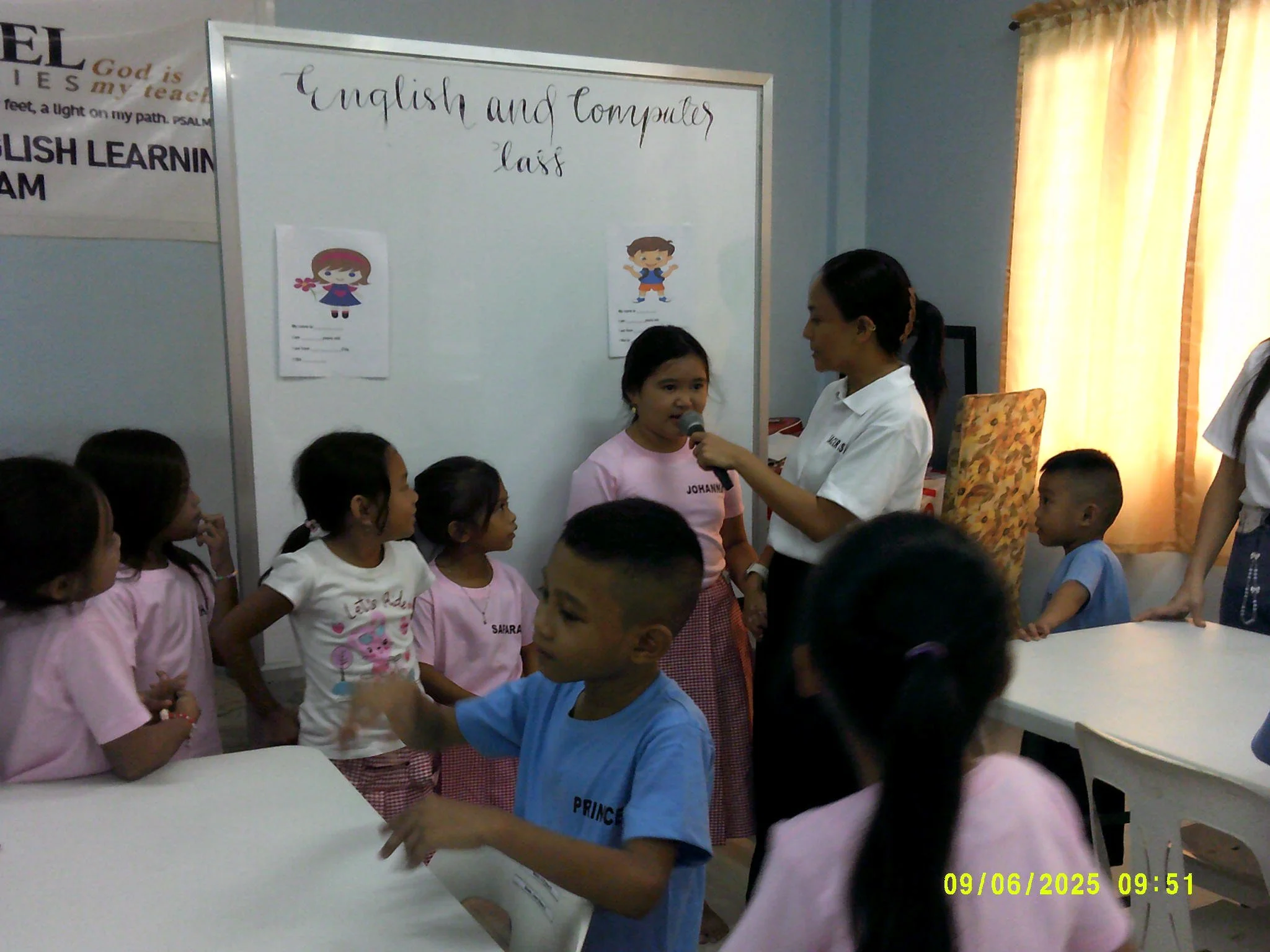 A classroom scene with children participating in an English and computer lesson. A girl is speaking into a microphone held by a teacher, surrounded by other children watching and interacting.
