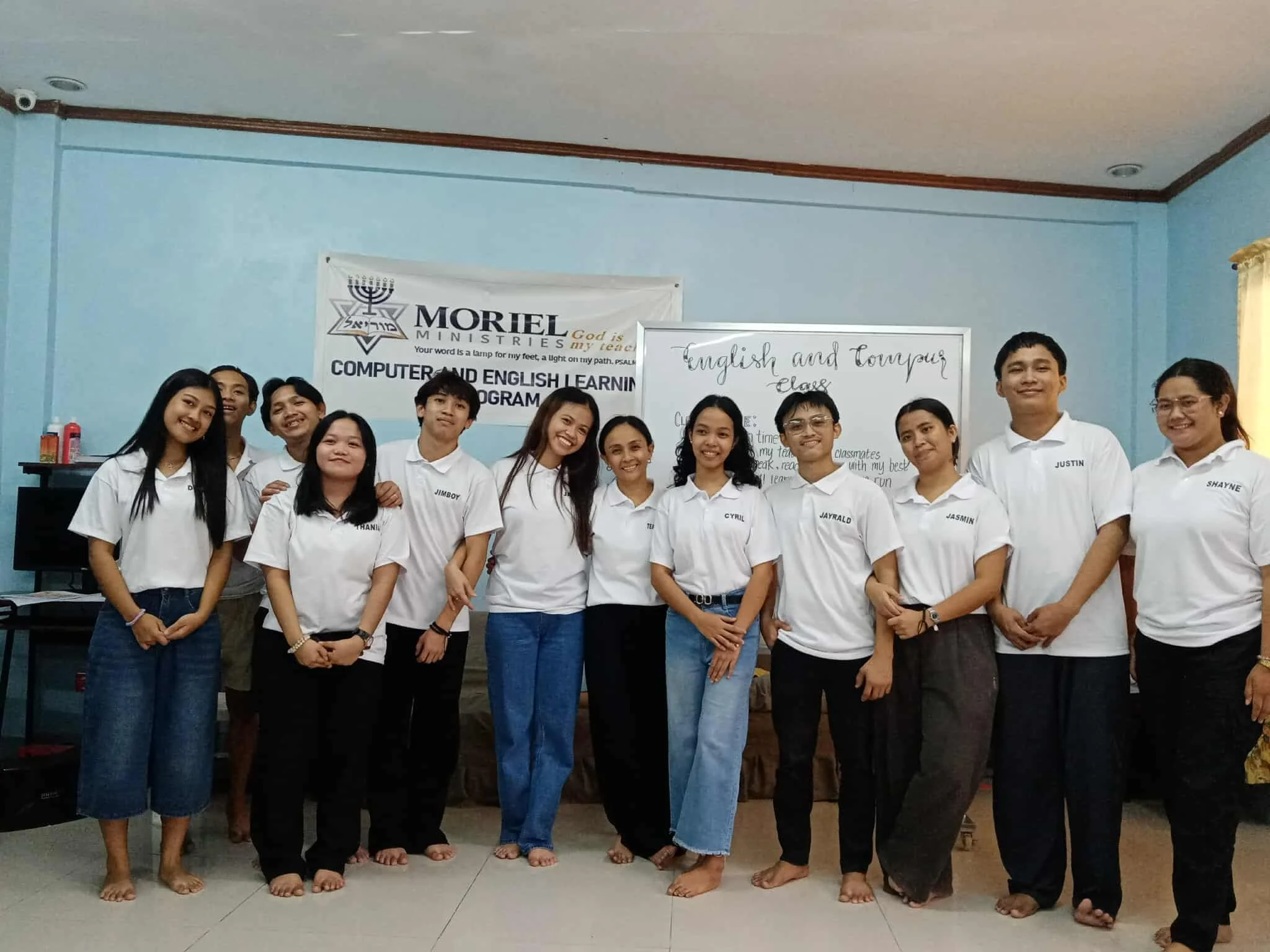 Group of students standing indoors for a photo, wearing white T-shirts with names, in front of a whiteboard and a banner about computer and English learning.