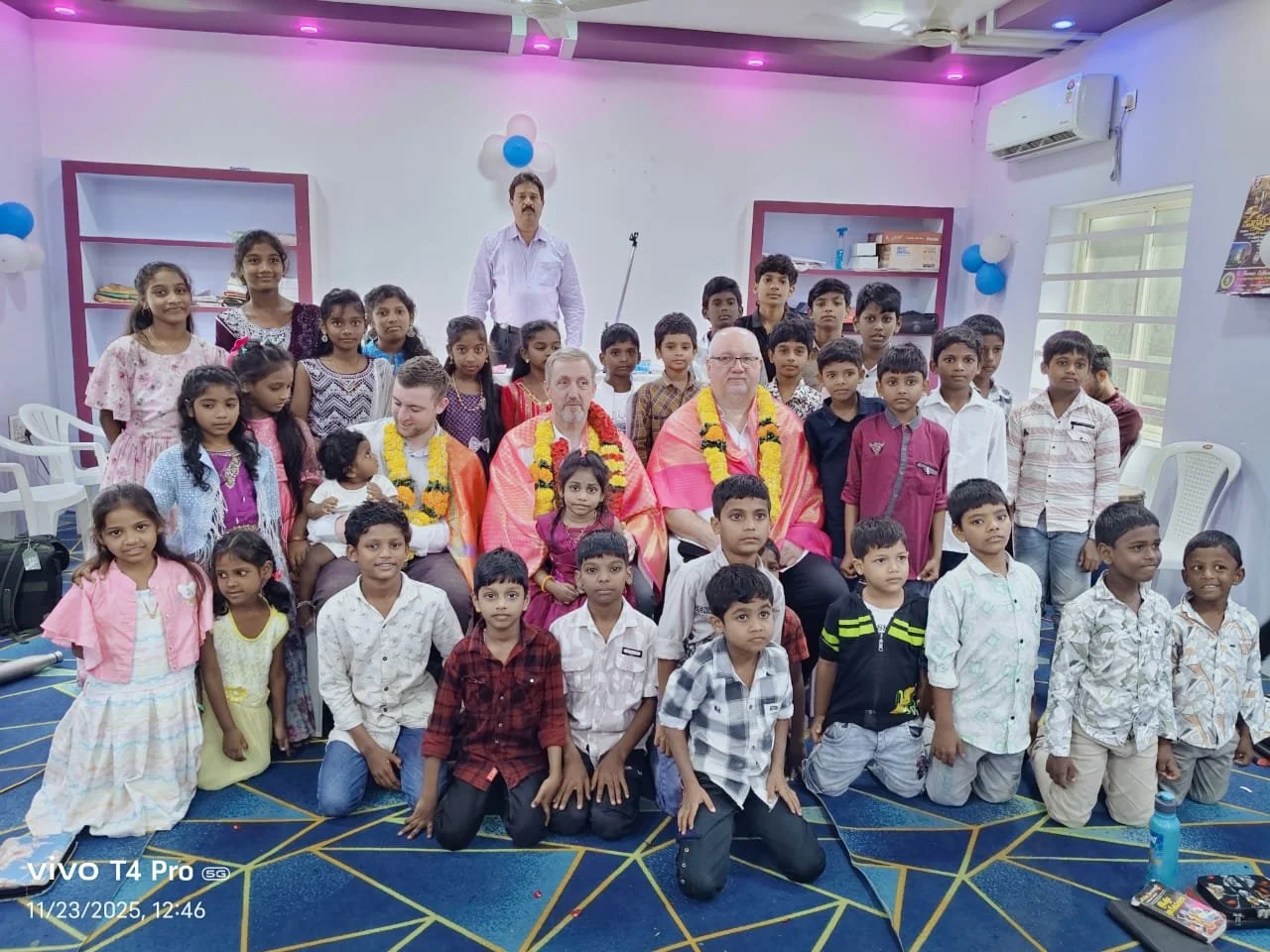 Group of children and adults gathered in a room, some wearing garlands, for a celebration or ceremony.