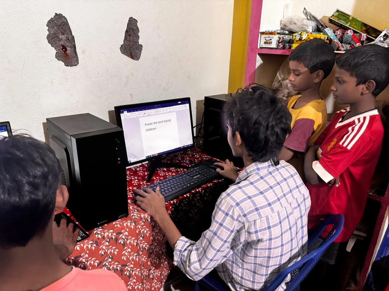 A group of children gathered around a computer monitor with an adult sitting at the desk, watching the screen. The monitor displays a text document with the words, 'Praise the lord moriel children.' The children are focused on the screen, standing and sitting in a small, cluttered room with snacks and boxes on shelves in the background.