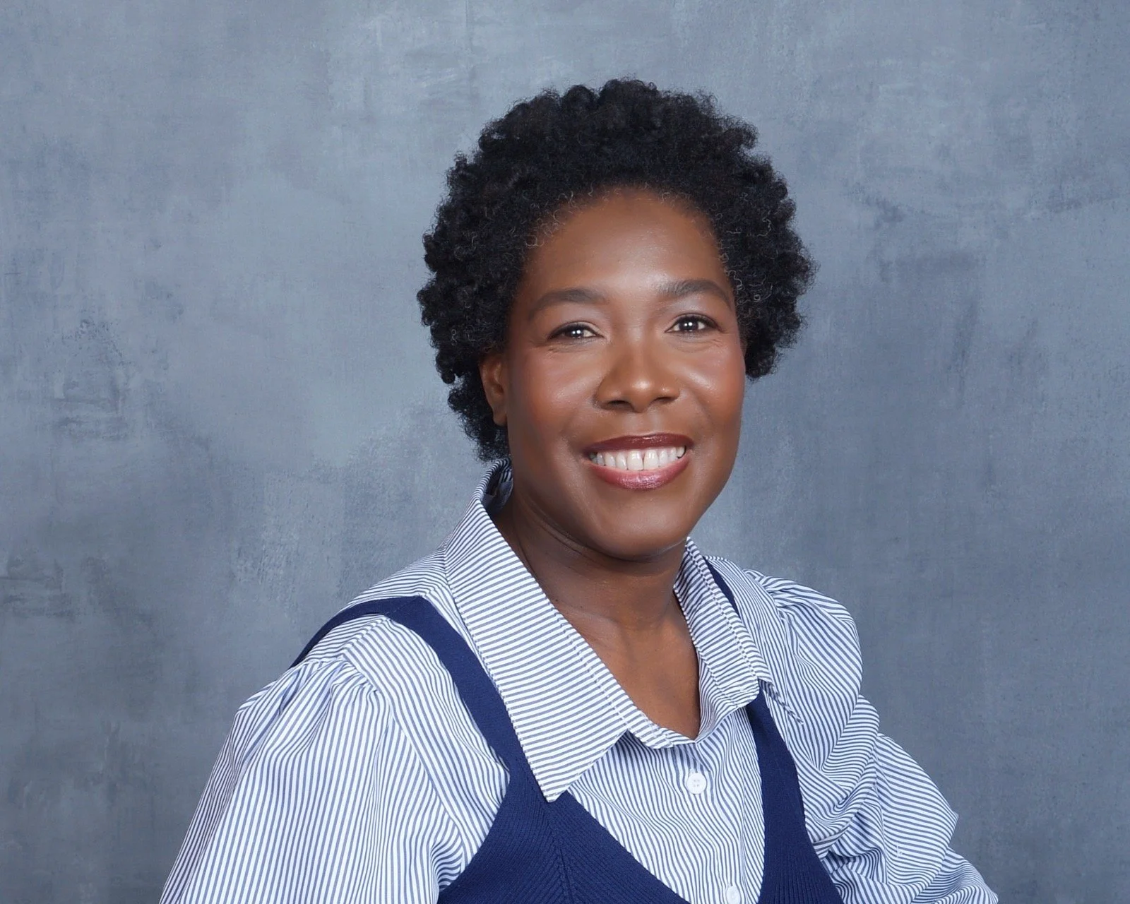 A woman with dark curly hair smiling, wearing a blue and white striped collared shirt and a navy blue sweater vest, against a gray textured background.