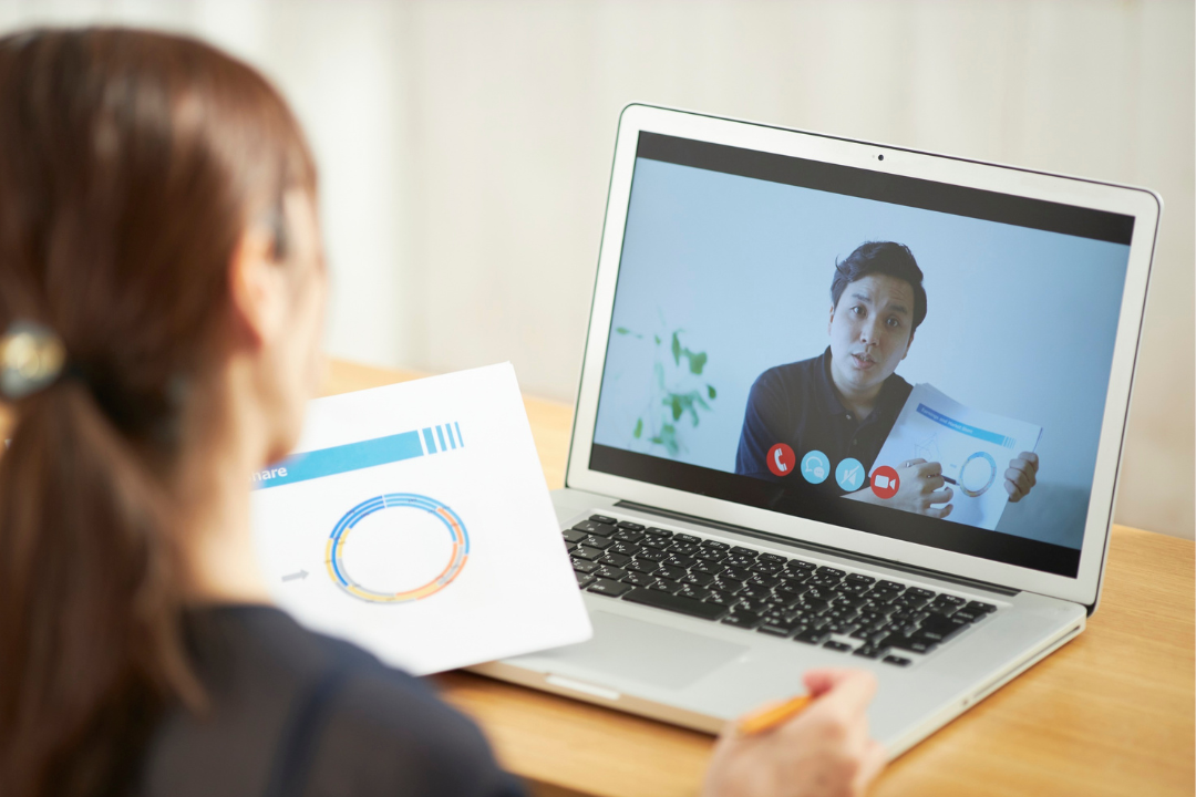 A woman participating in a video call on her laptop, holding a printed chart with a circular graph, while the man on the screen holds a document and appears to be explaining something.