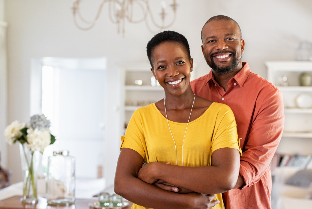 A smiling African American couple stands close together in their living room, with the woman in a yellow shirt and the man in an orange shirt, looking at the camera.