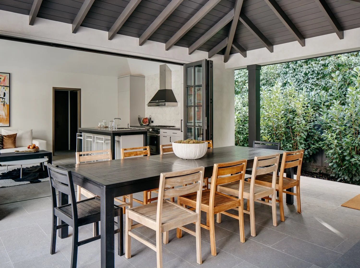 An outdoor covered dining pavilion furnished with a long dark wooden table, chairs in various wood tones, with an open view of the surrounding landscaping; a kitchen and sitting area are visible in the background.