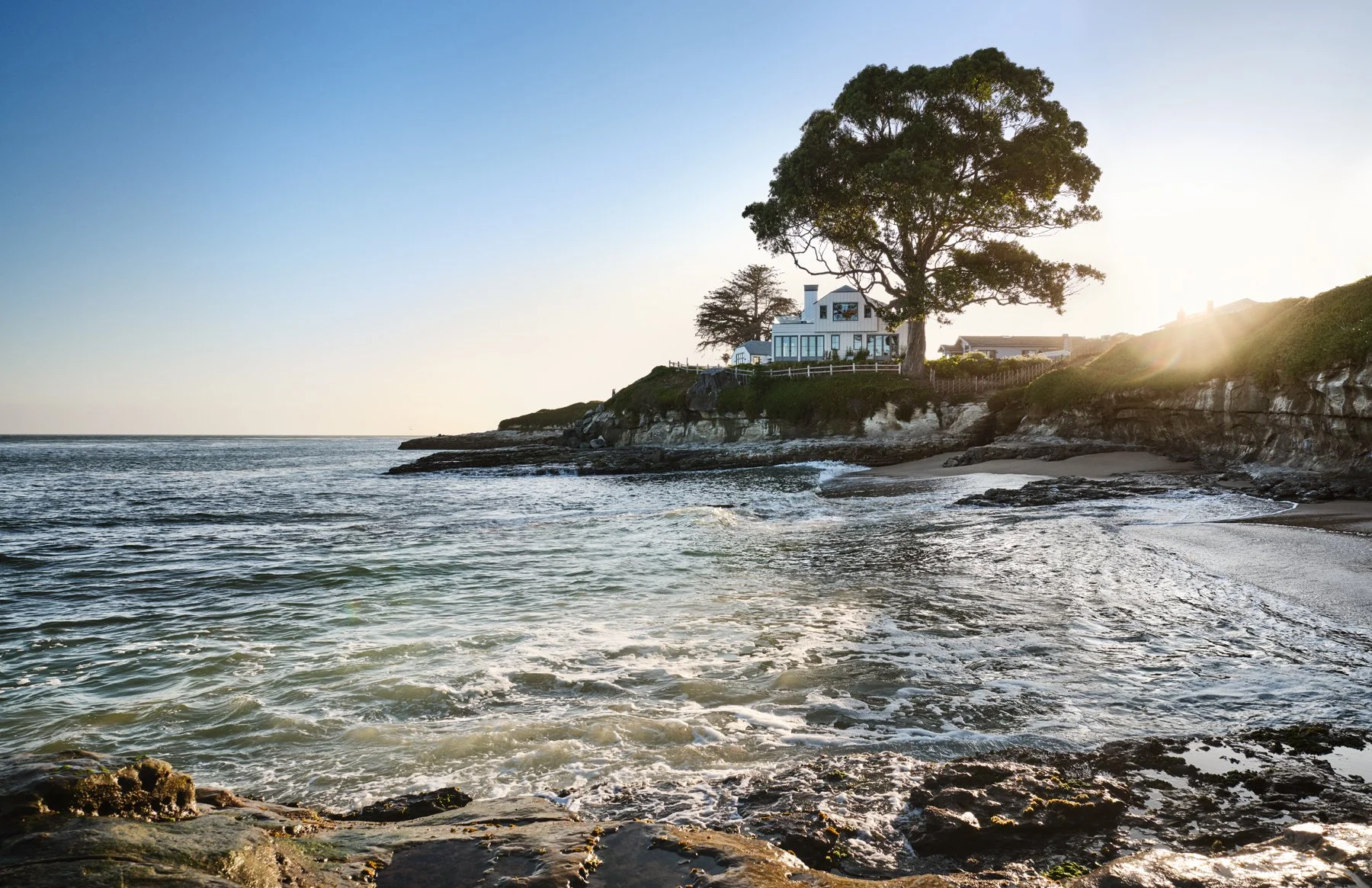 A pale house sits on a rocky shoreline above the water; the house is partly screened by a mature tree.