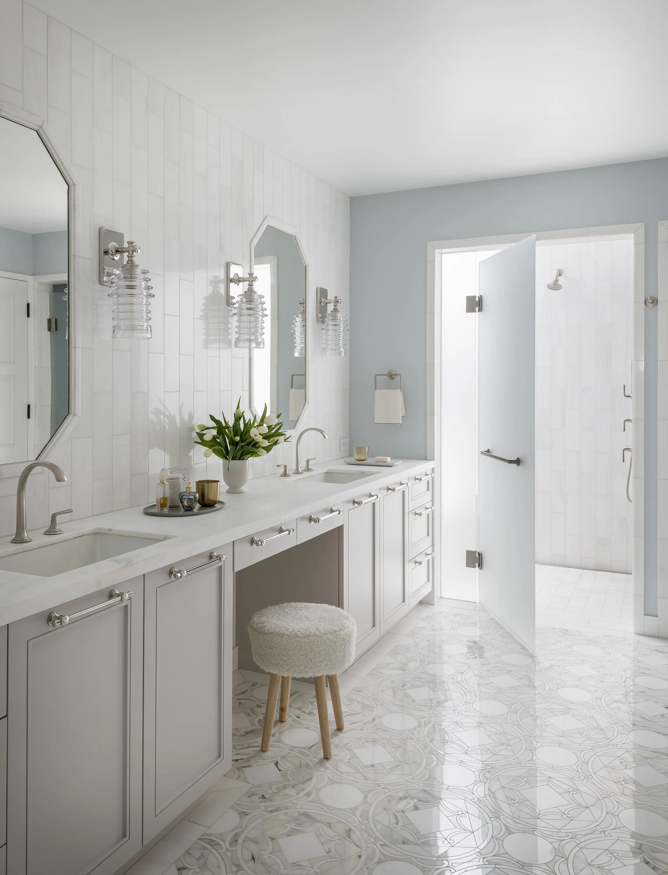 Clean, bright bathroom vanity area featuring pale painted cabinetry, two octagonal mirrors, and two sinks in a long white stone countertop, with an open shower door on the far wall.