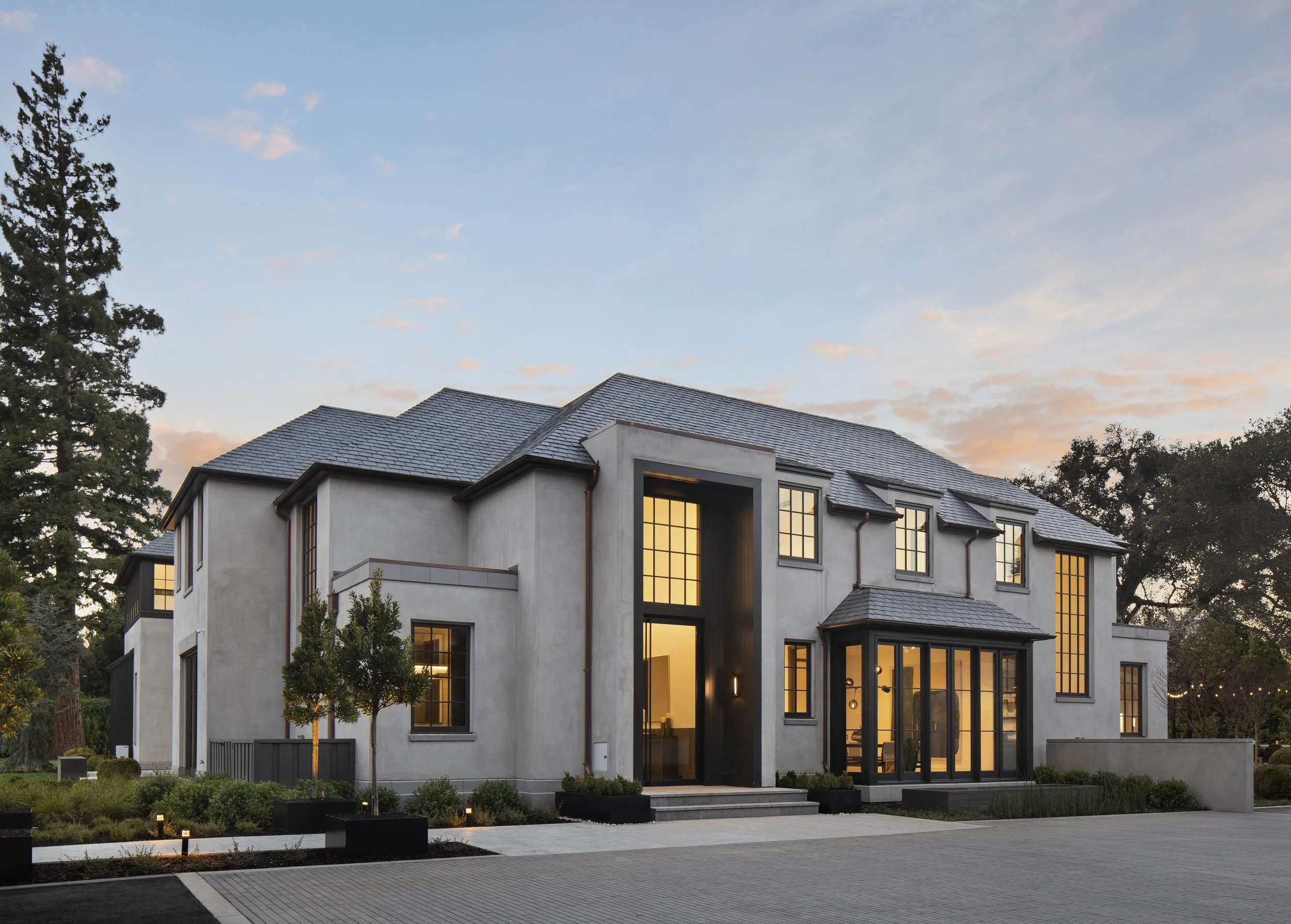 An exterior twilight view of a two-story residence featuring a multi-gable roof, tall ground-floor windows, and stone facade, with a paved motor court and mature trees in the background.