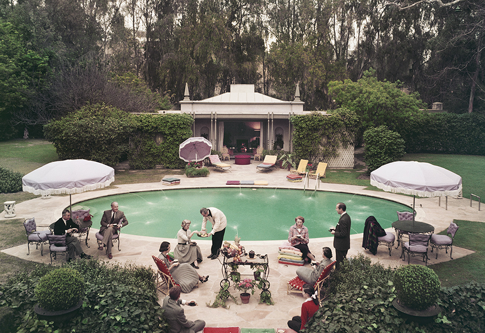 A group of elegantly dressed people gather around a swimming pool in the landscaped grounds of a house.