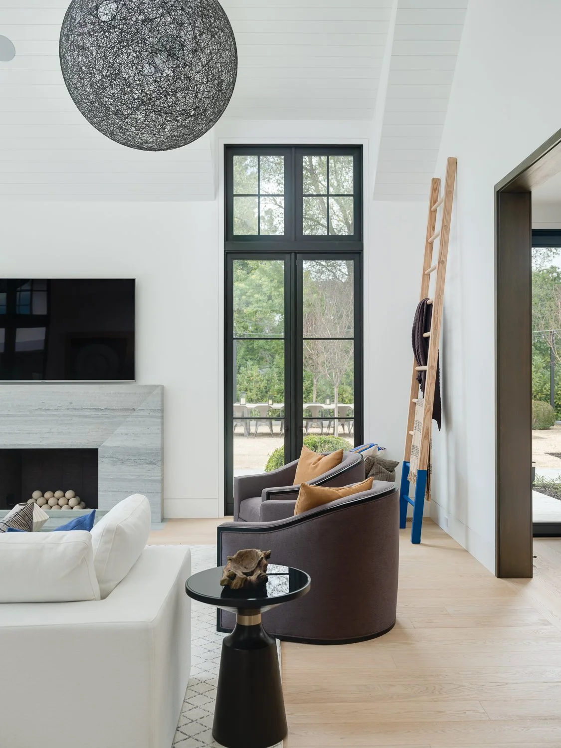 A living room area featuring a light stone fireplace, white walls and ceiling, two dark upholstered chairs, a white sofa, and floor-to-ceiling glass French doors with dark trim leading to the exterior.