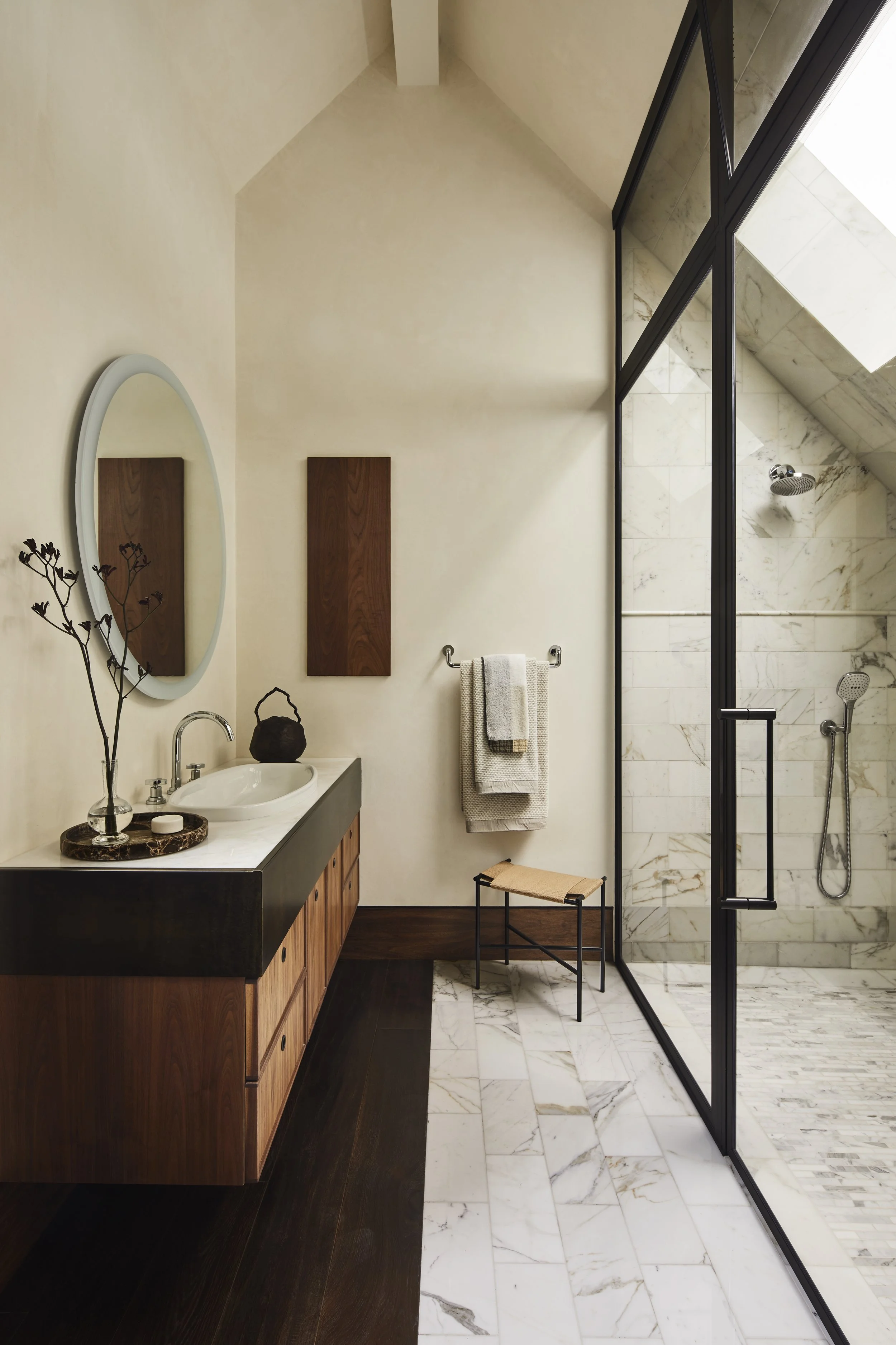 Bright, airy bathroom featuring a vaulted ceiling with a white wooden beam, a floating wooden vanity with a marble countertop and basin sink, a round illuminated mirror, and a large black-framed glass wall leading into a marble-tiled walk-in shower.