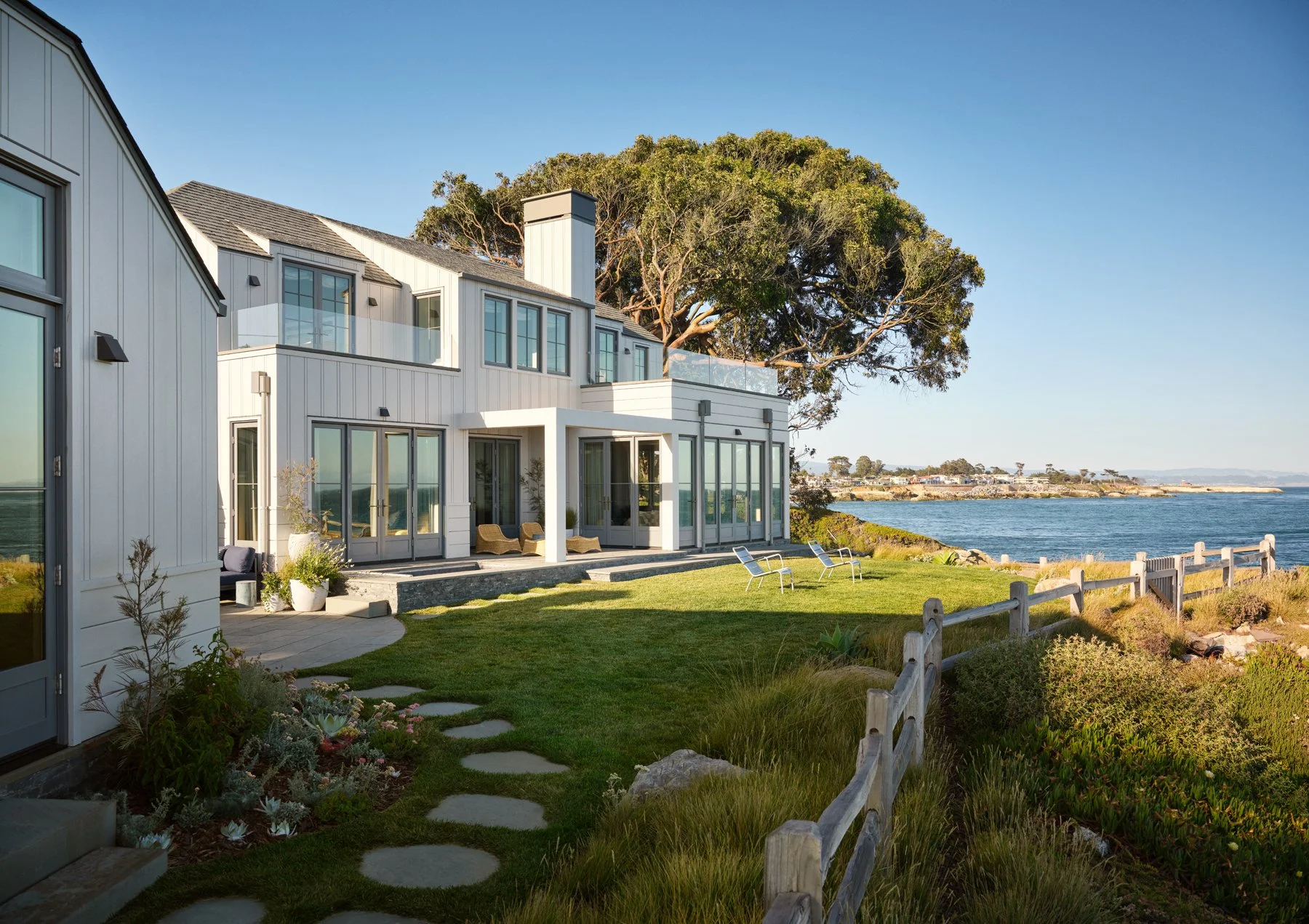 An exterior view of the two-level house facade featuring glass doors opening to a stone patio and lawn overlooking the ocean.