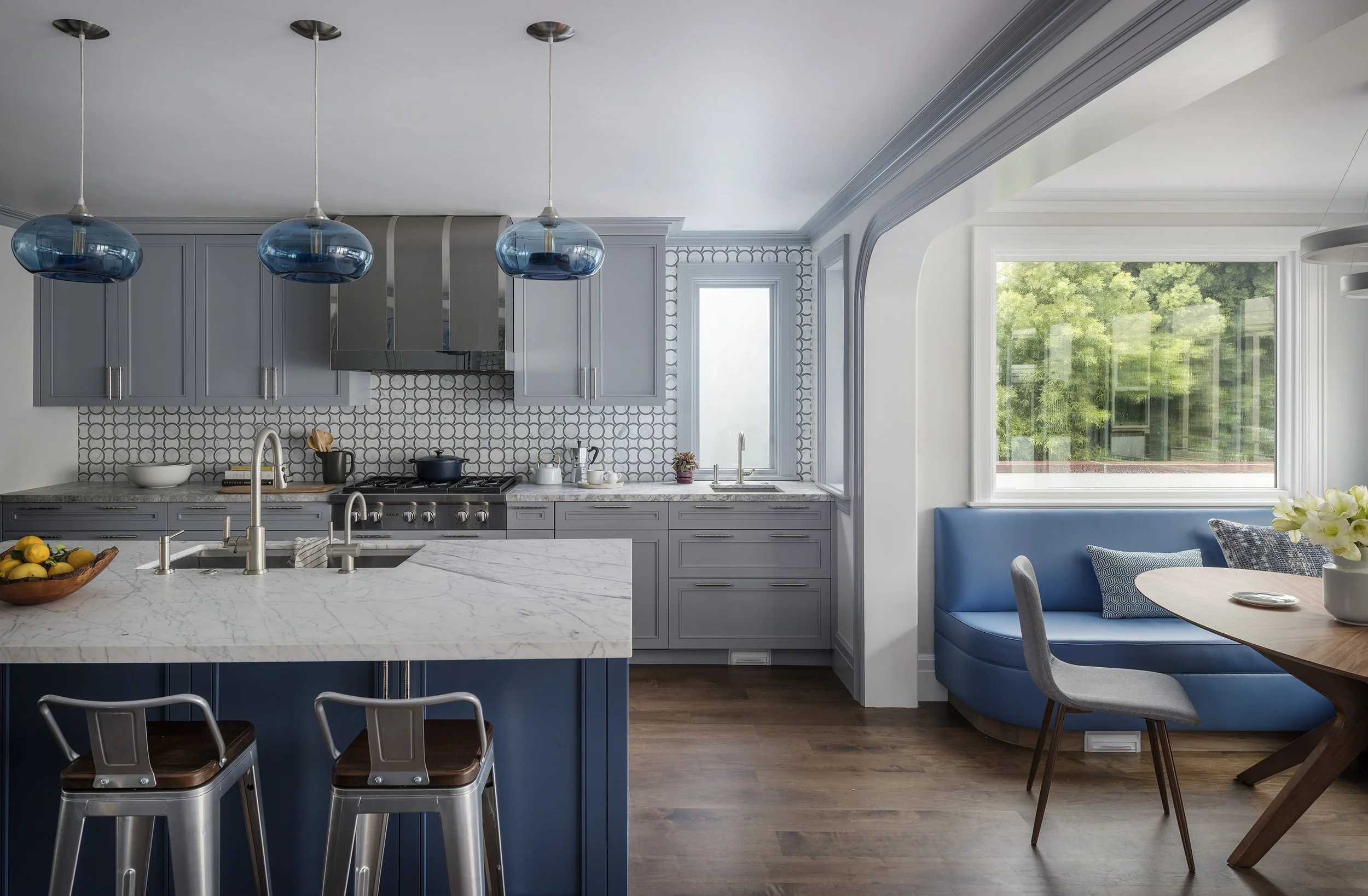 Modern kitchen with dark blue cabinetry on the island, pale blue-grey cabinets on the far wall, a stainless steel range, and a dining area with built-in blue upholstered seating.