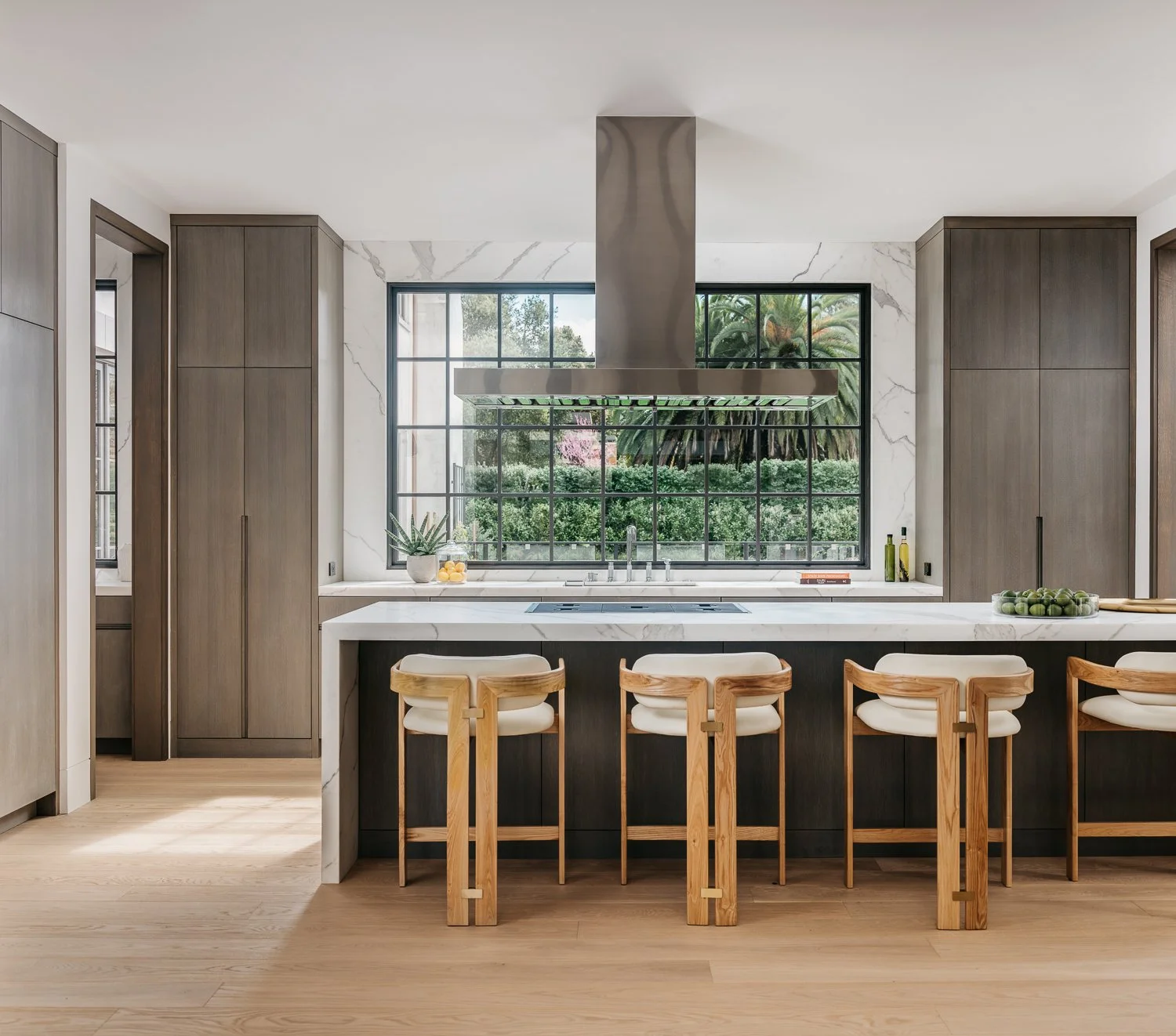 A kitchen interior featuring a central island with wood bar stools, white perimeter countertops, dark cabinetry, and large gridded window looking out to greenery on the far wall.