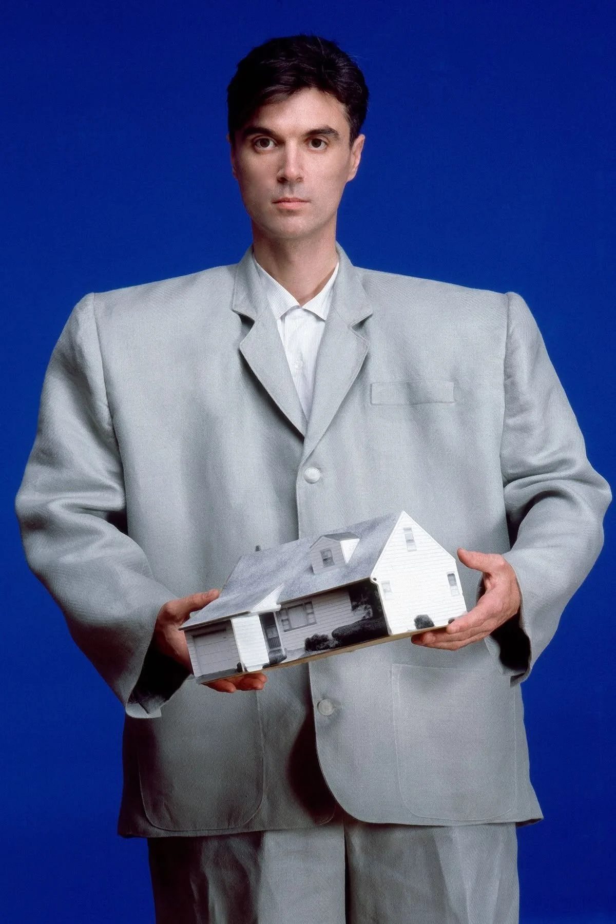 A man in a pale grey suit holds a small model of a house against a vivid blue background.