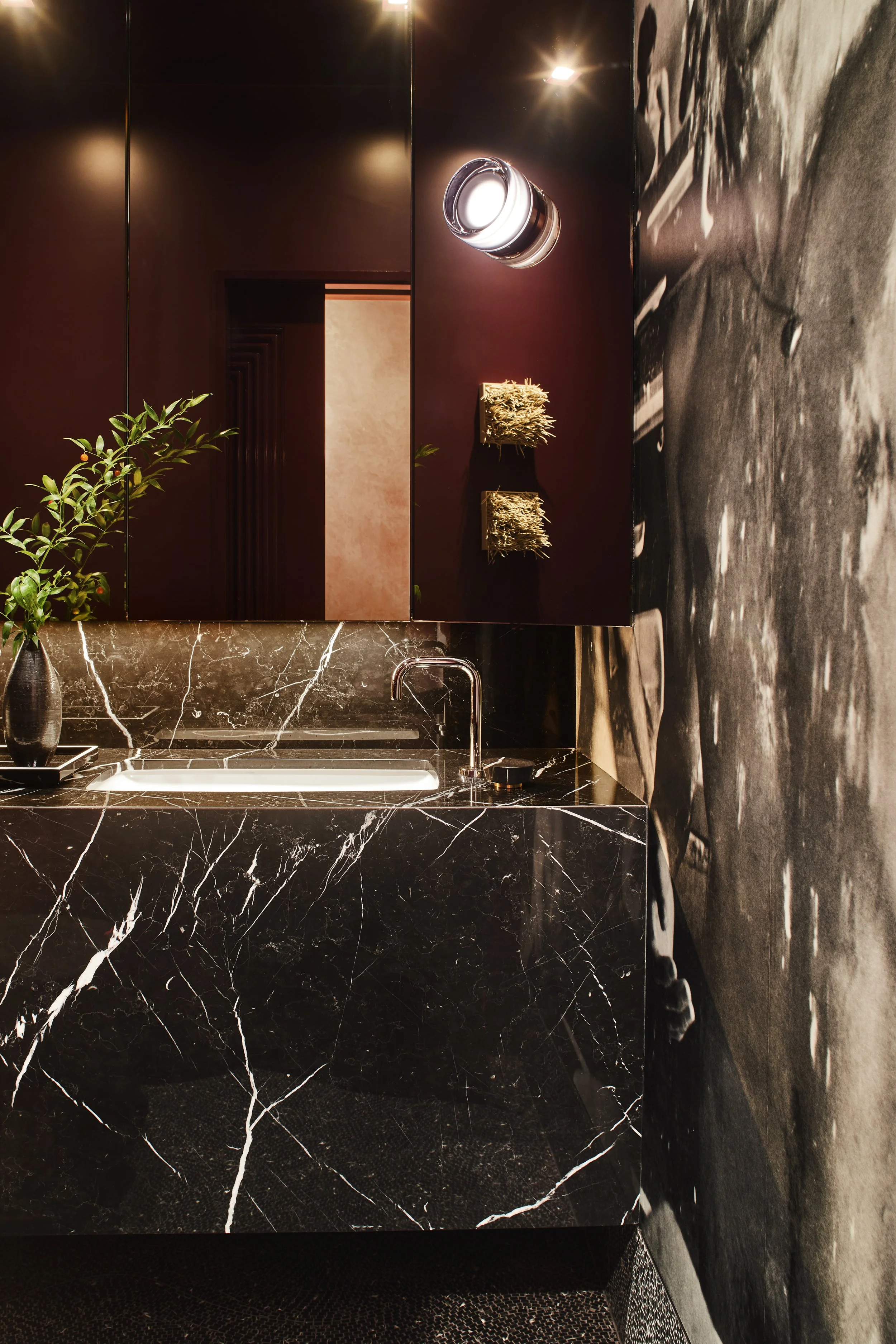 Close-up view of the dark stone powder room vanity, with dark red walls, a circular sconce, and photographic black-and-white wallpaper.