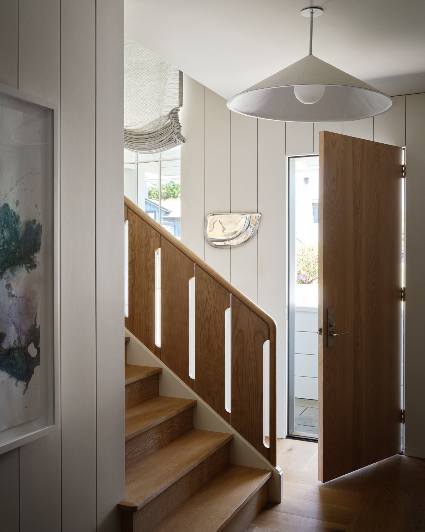 An interior staircase with wooden treads, a handrail featuring vertical cutouts, and tall vertical windows looking out toward exterior greenery, alongside an open wooden entry door.