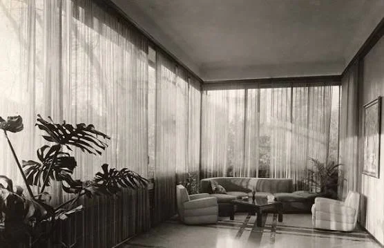A light-colored curved sofa sits in a sunlit room surrounded by tall sheer curtains and tropical plants.