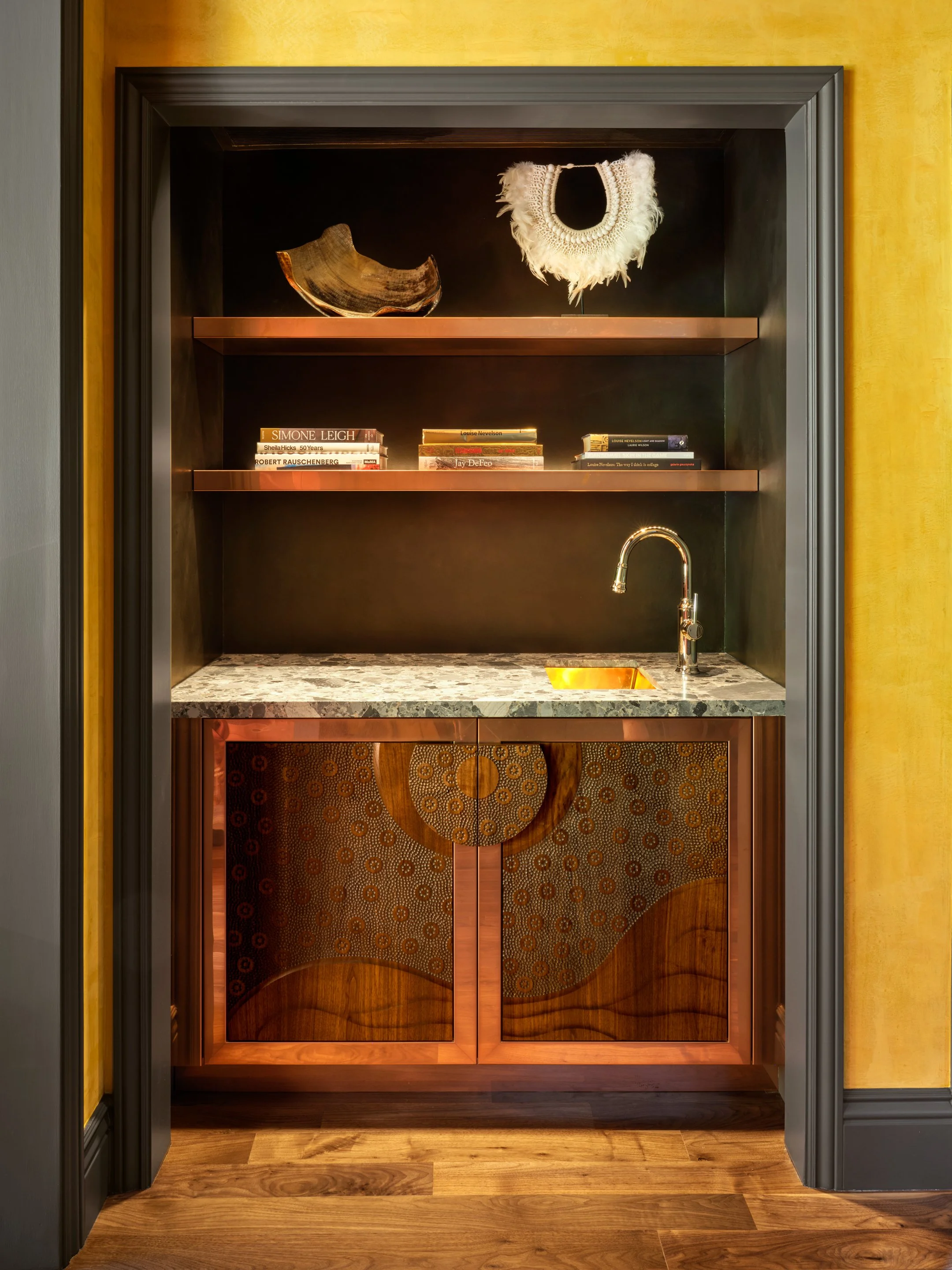 Recessed built-in bar and shelving featuring rich wood tones, a stone countertop, dark walls and trim, and a brass sink, with warm yellow plaster walls visible in the foreground.