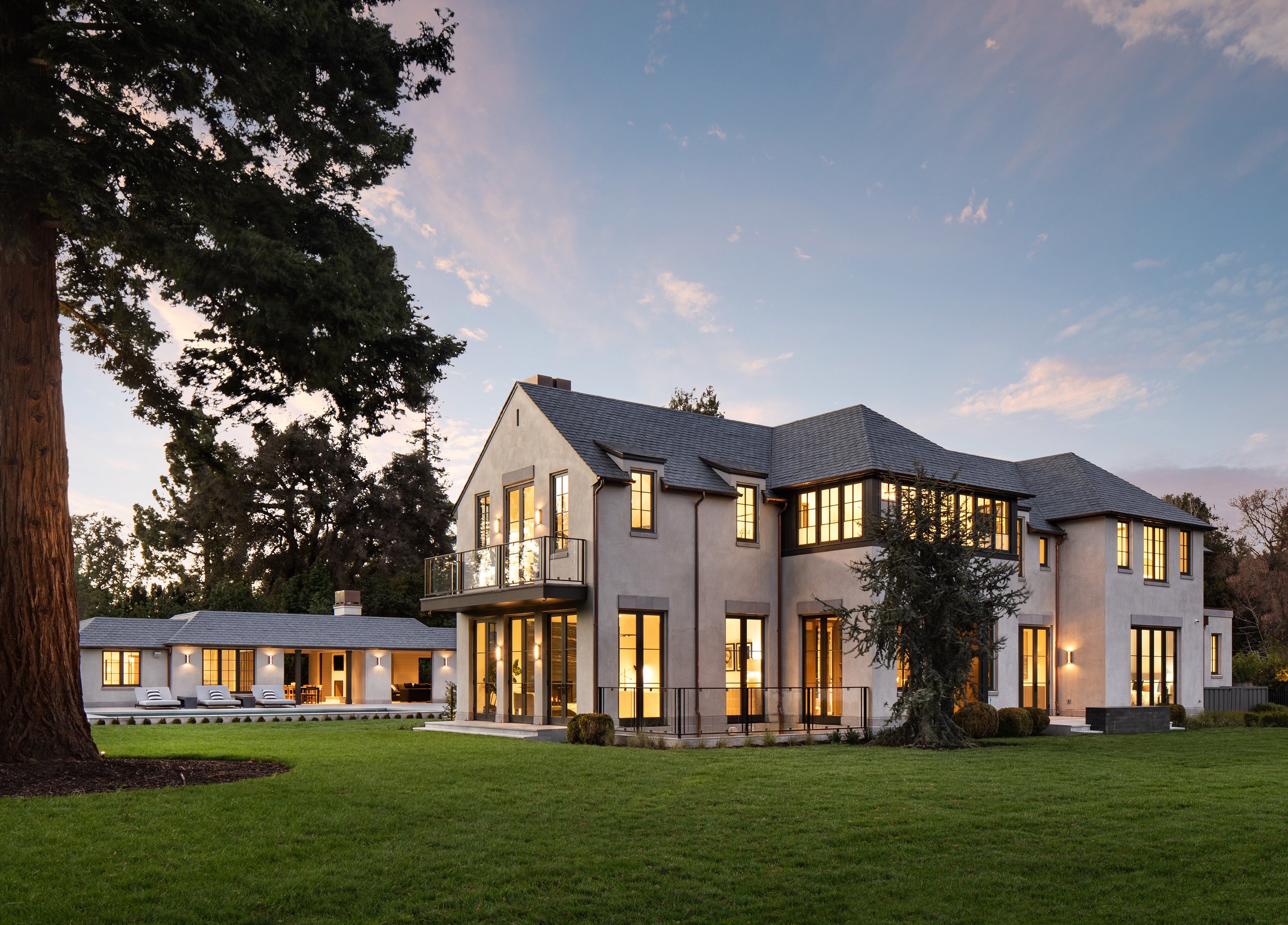 An exterior twilight view of a two-story residence featuring a multi-gable roof, tall ground-floor windows, and stone facade, with a manicured lawn and mature evergreen tree.