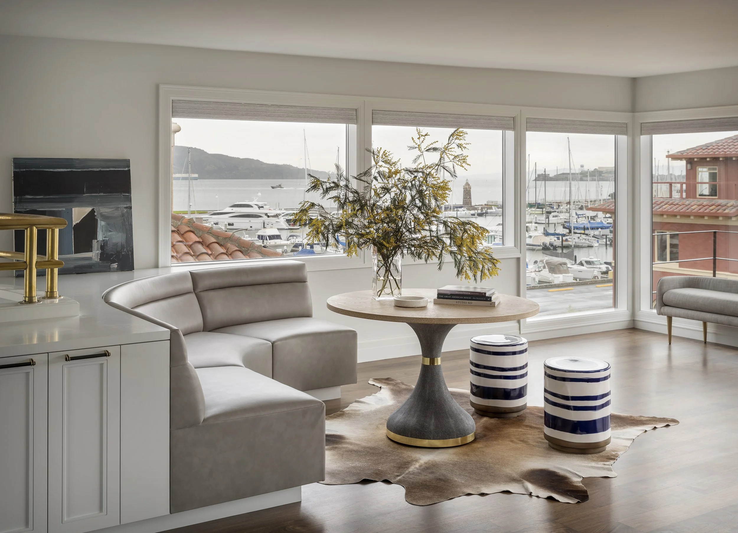 Bright, modern dining area featuring a curved built-in banquette with neutral upholstery, a round wooden dining table, two cylindrical stools, and large windows overlooking a marina.