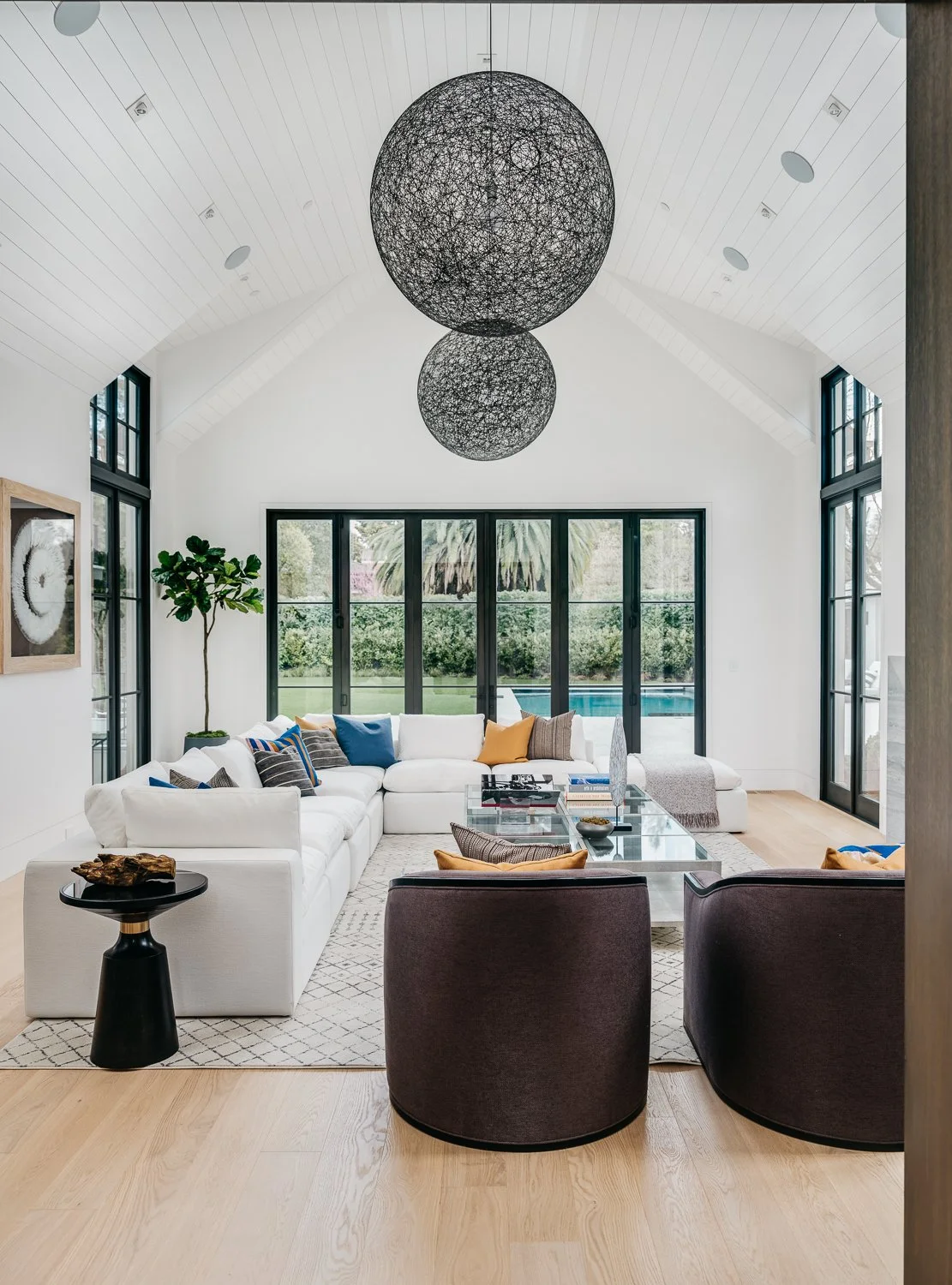 A living room featuring a large sectional white sofa, white walls and ceiling, two dark upholstered chairs, and a wall of floor-to-ceiling glass doors with dark trim leading to the pool area.