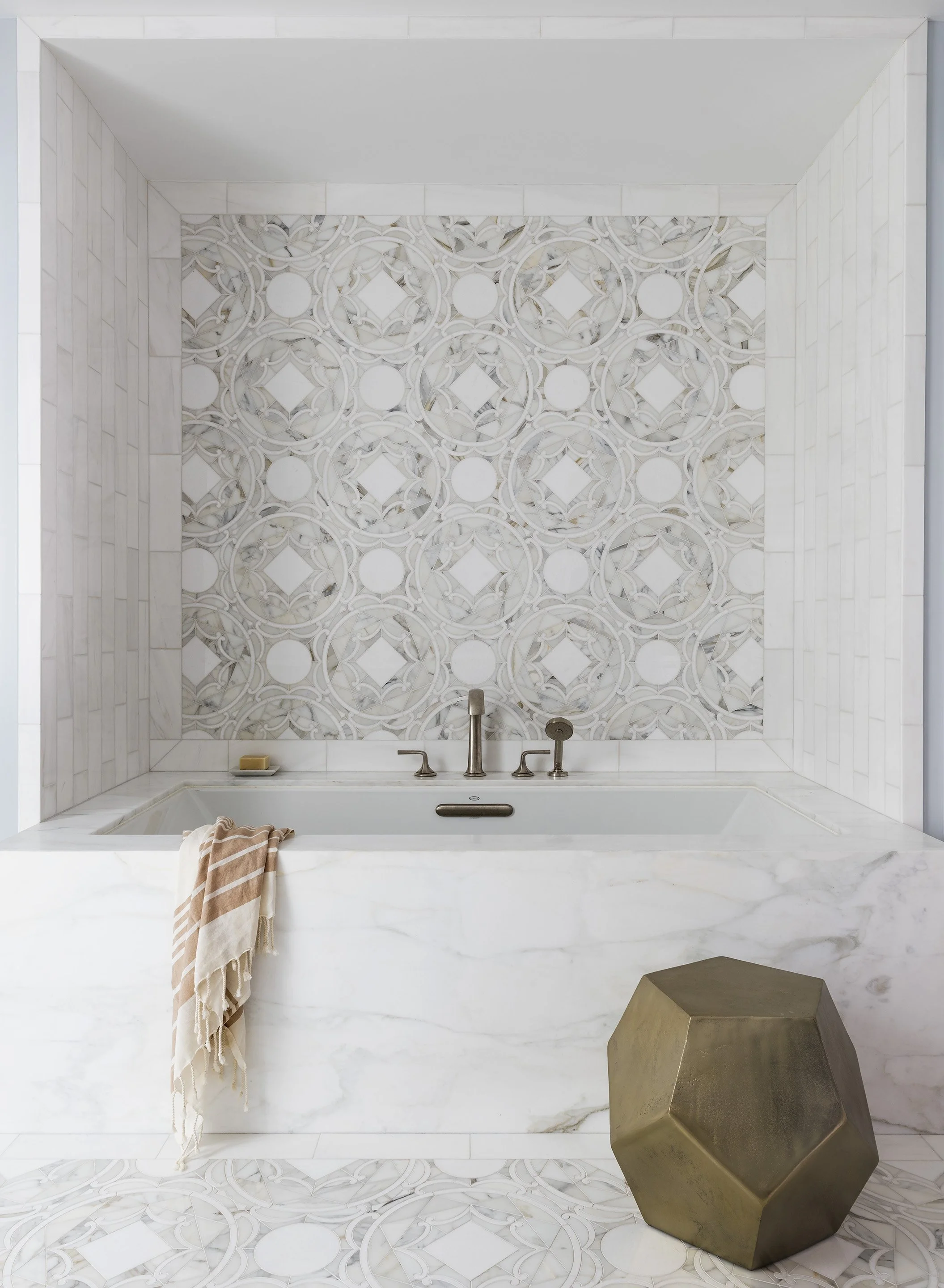Interior view of a bathroom featuring a soaking tub against a decorative geometric tiled feature wall, with warm natural light.