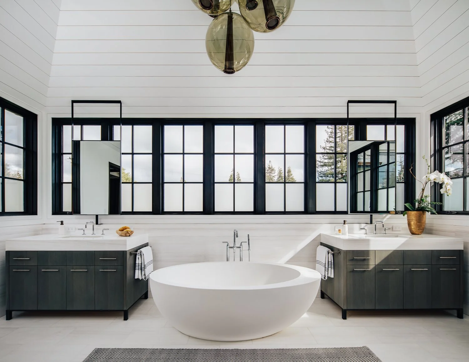 A brightly lit bathroom containing a freestanding white soaking tub, dark cabinetry with dual sinks, and a large multi-pane window with exterior views.