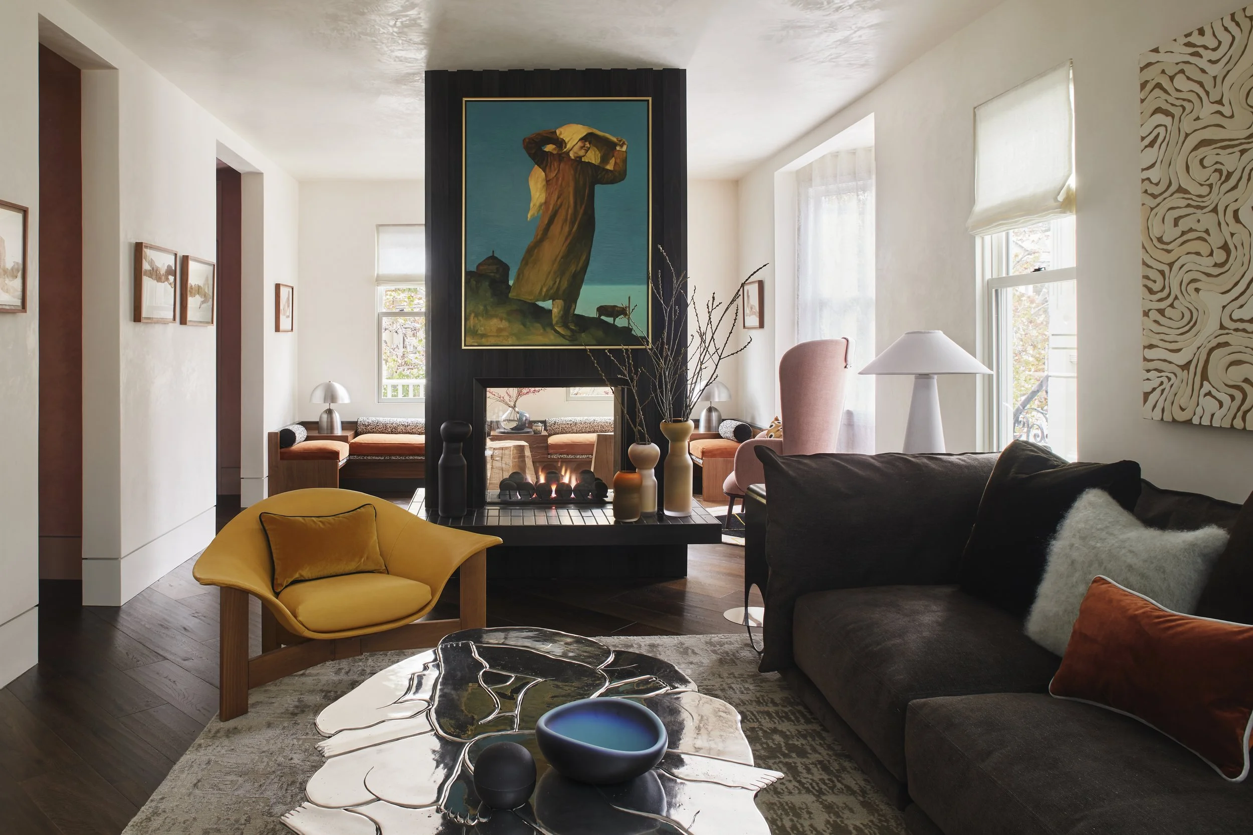 A black-framed double-sided fireplace divides two seating areas, with a large painting above it. A mustard-yellow lounge chair and dark brown sofa face a sculptural reflective coffee table on a patterned rug.