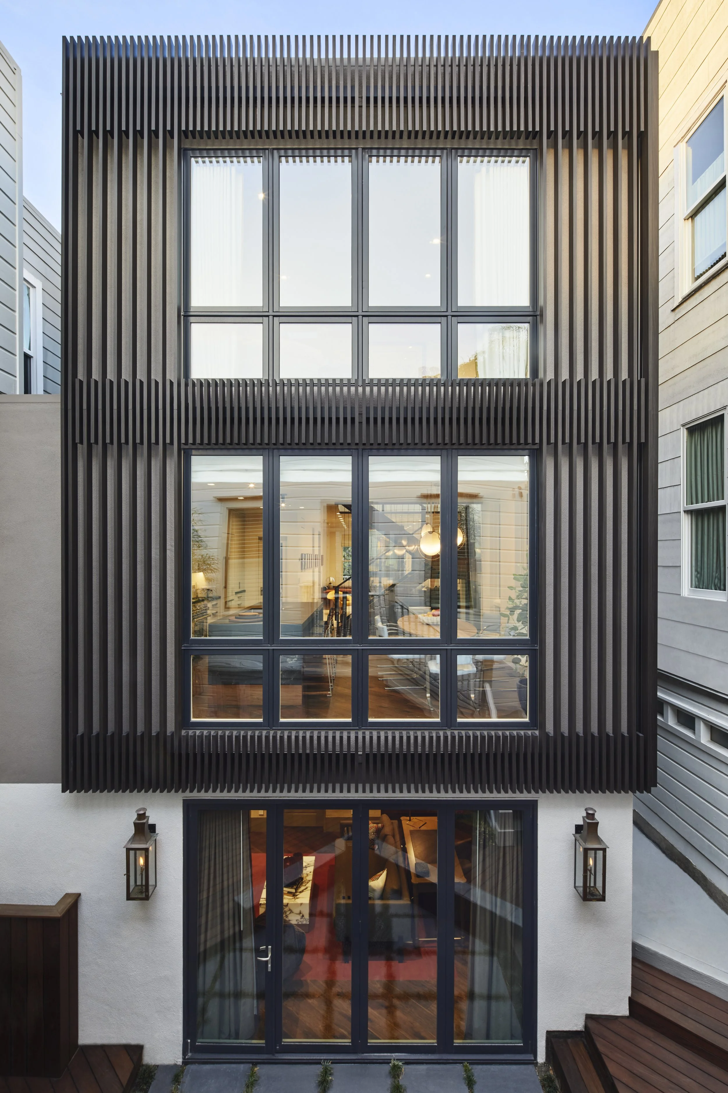 Rear exterior view of a multi-story home featuring dark vertical louvers covering the upper two stories, large multi-pane windows, and white stucco walls at the ground level with glass double doors opening onto a rear yard.