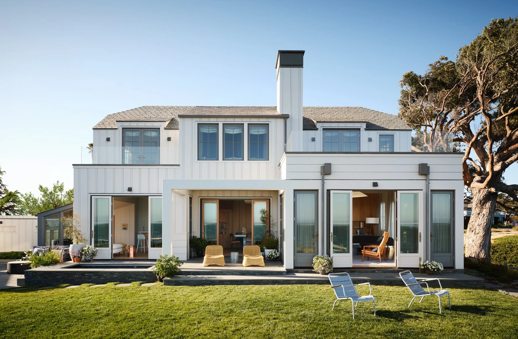 An exterior view of the two-level house facade featuring three sets of glass doors connecting different interior spaces to a stone patio and lawn in the foreground.