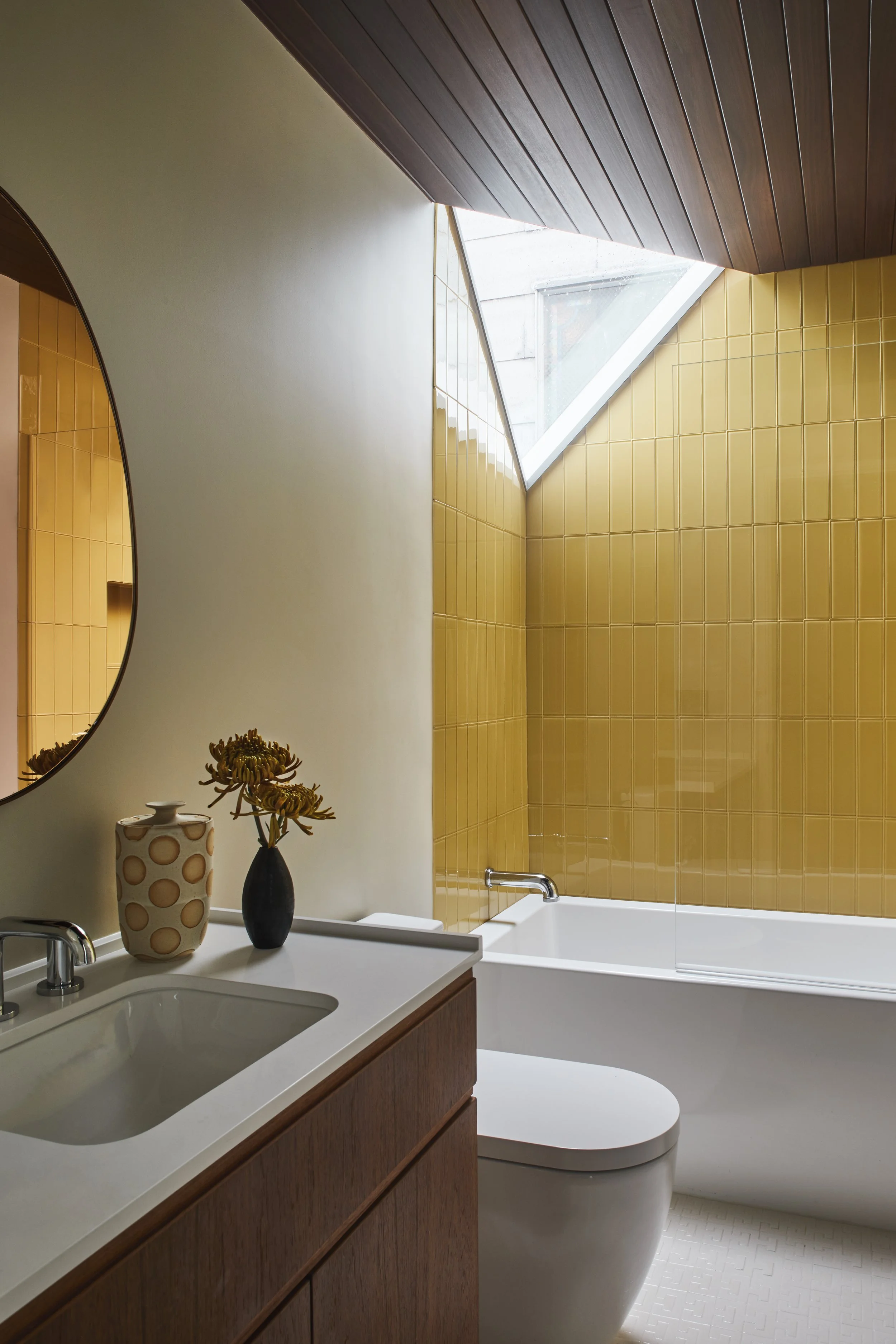 Bright, modern bathroom featuring warm mustard-yellow vertical tile walls on one side, a striking triangular corner window above the white tub, and a dark wood vanity with a white countertop and a round mirror on the left.