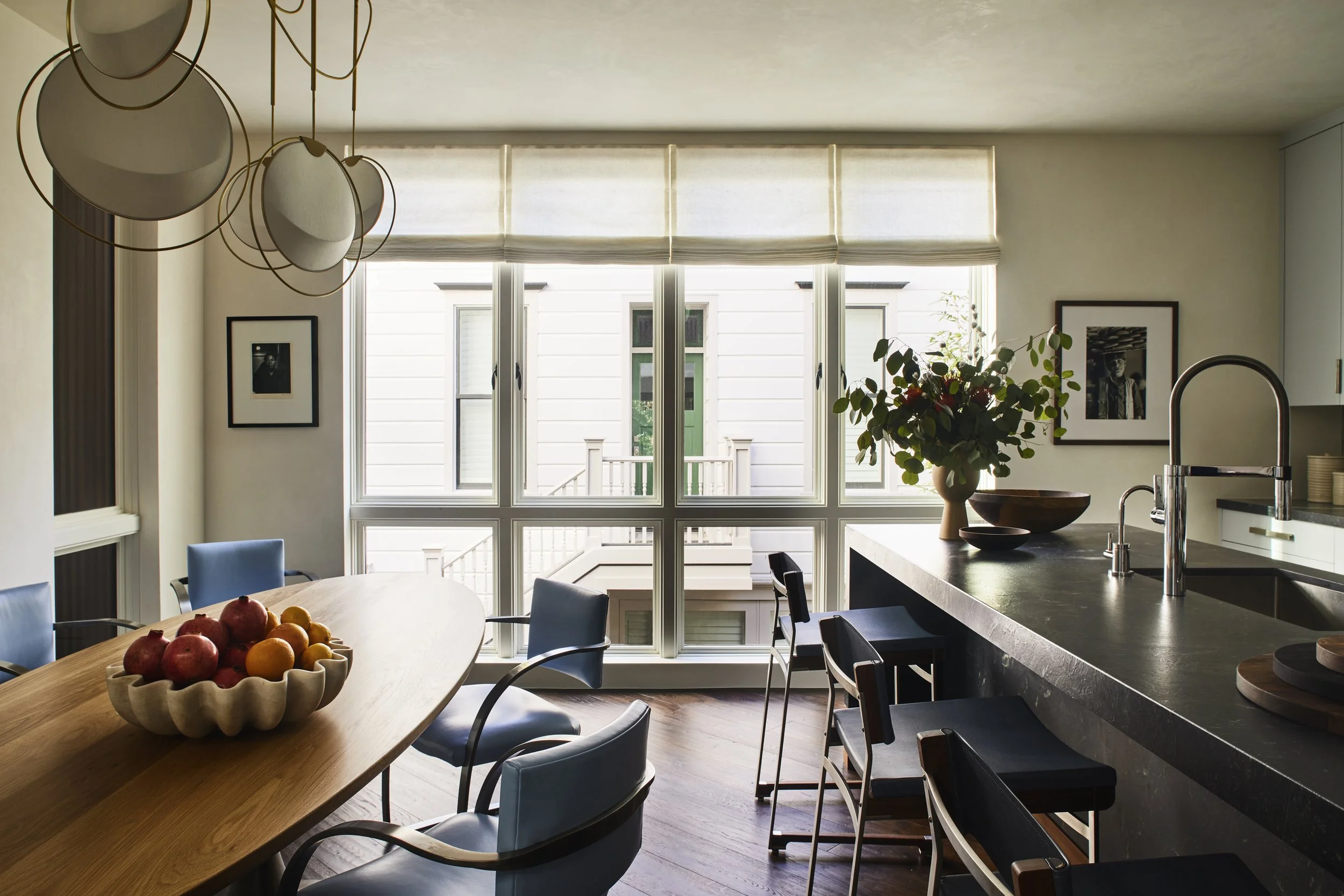 Kitchen and dining area, featuring a long oval wood dining table wth light blue upholstered chairs, a dark kitchen island, and a wall of floor-to-ceiling windows.