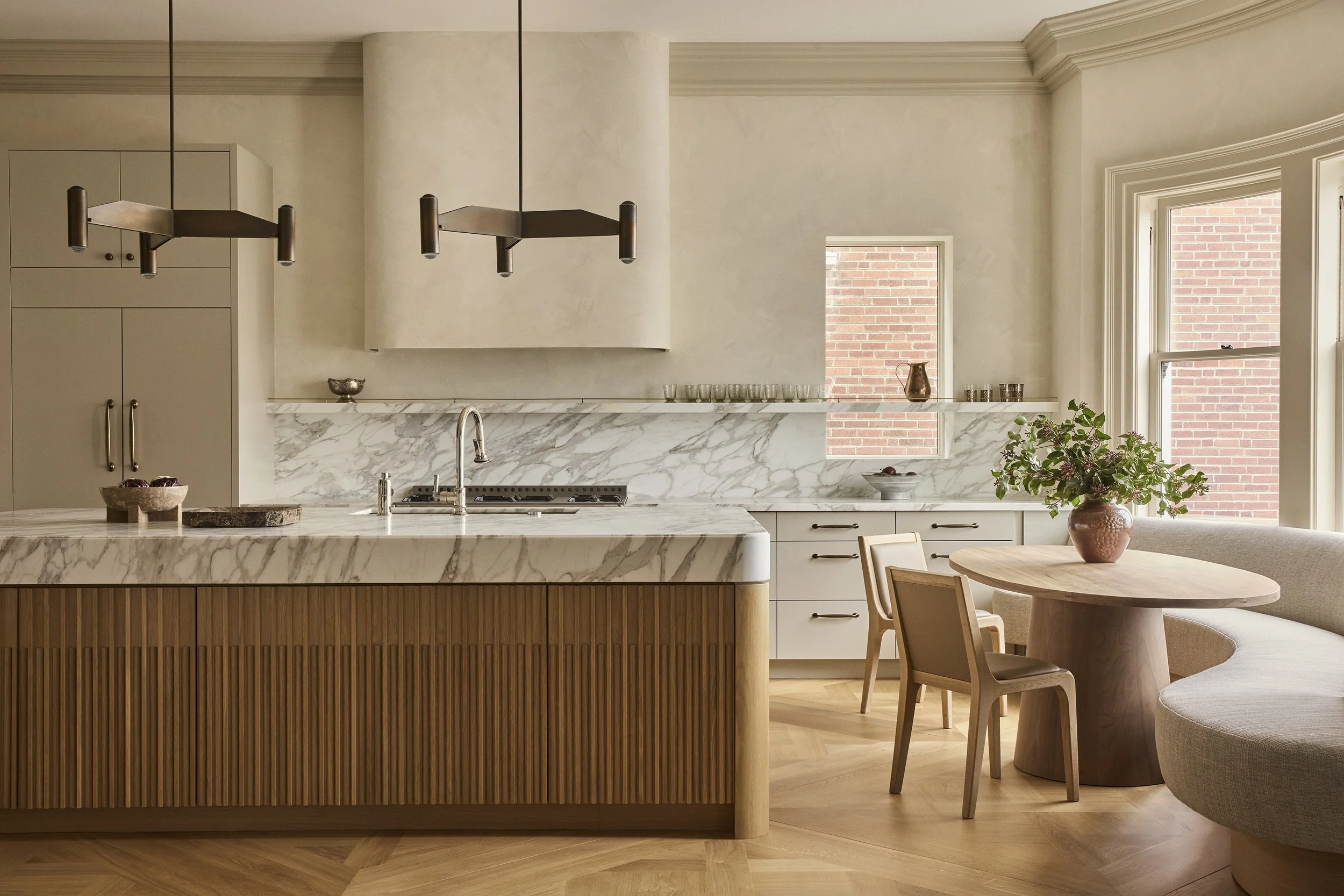 A fluted wood kitchen island with a veined marble countertop faces pale cabinetry and a matching marble backsplash. A round wood table and curved upholstered banquette sit beside tall windows.