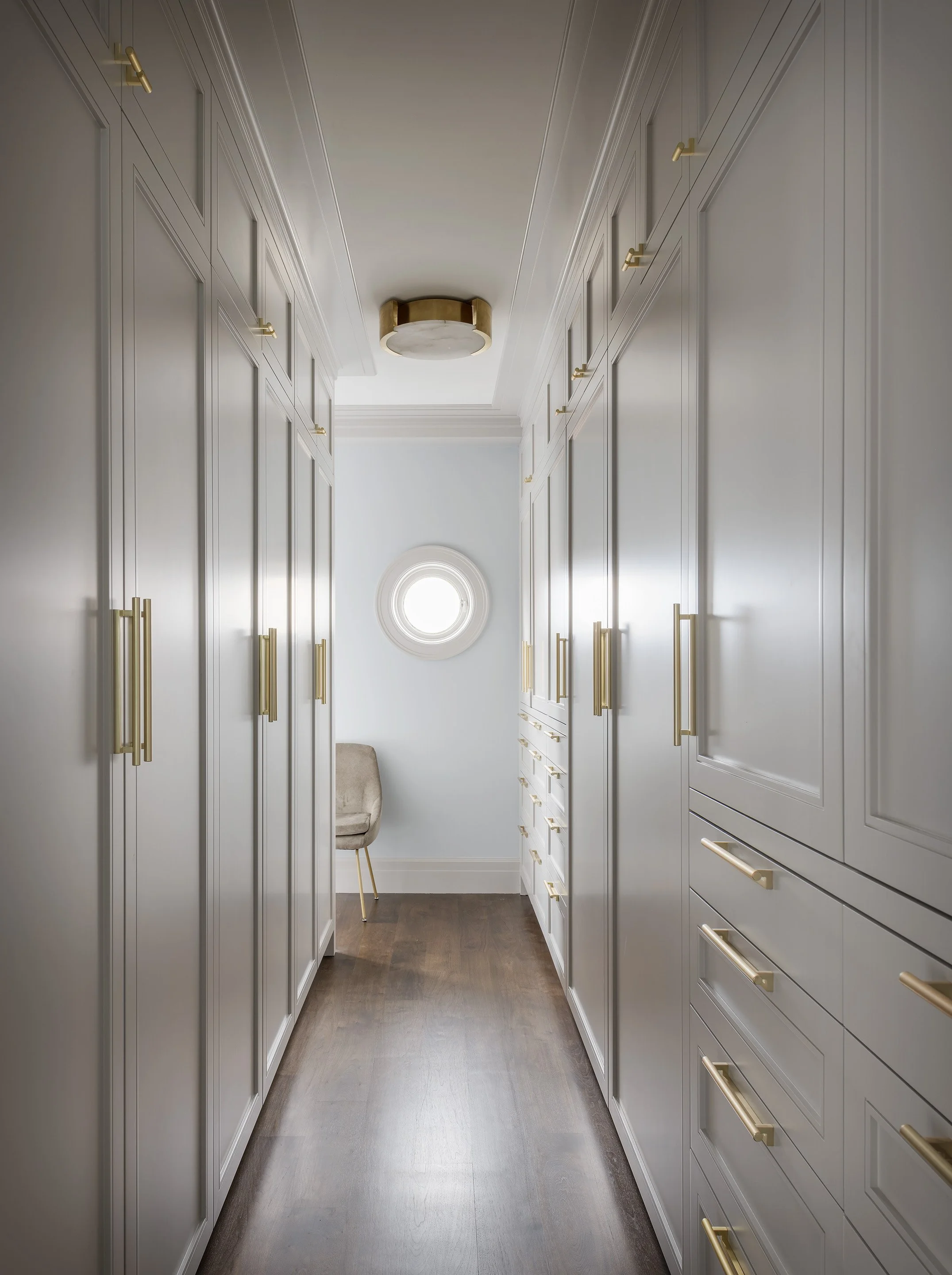 Long interior view of a storage area with built-in floor-to-ceiling cabinetry, minimalist brass pulls, and a circular accent window on the far wall.