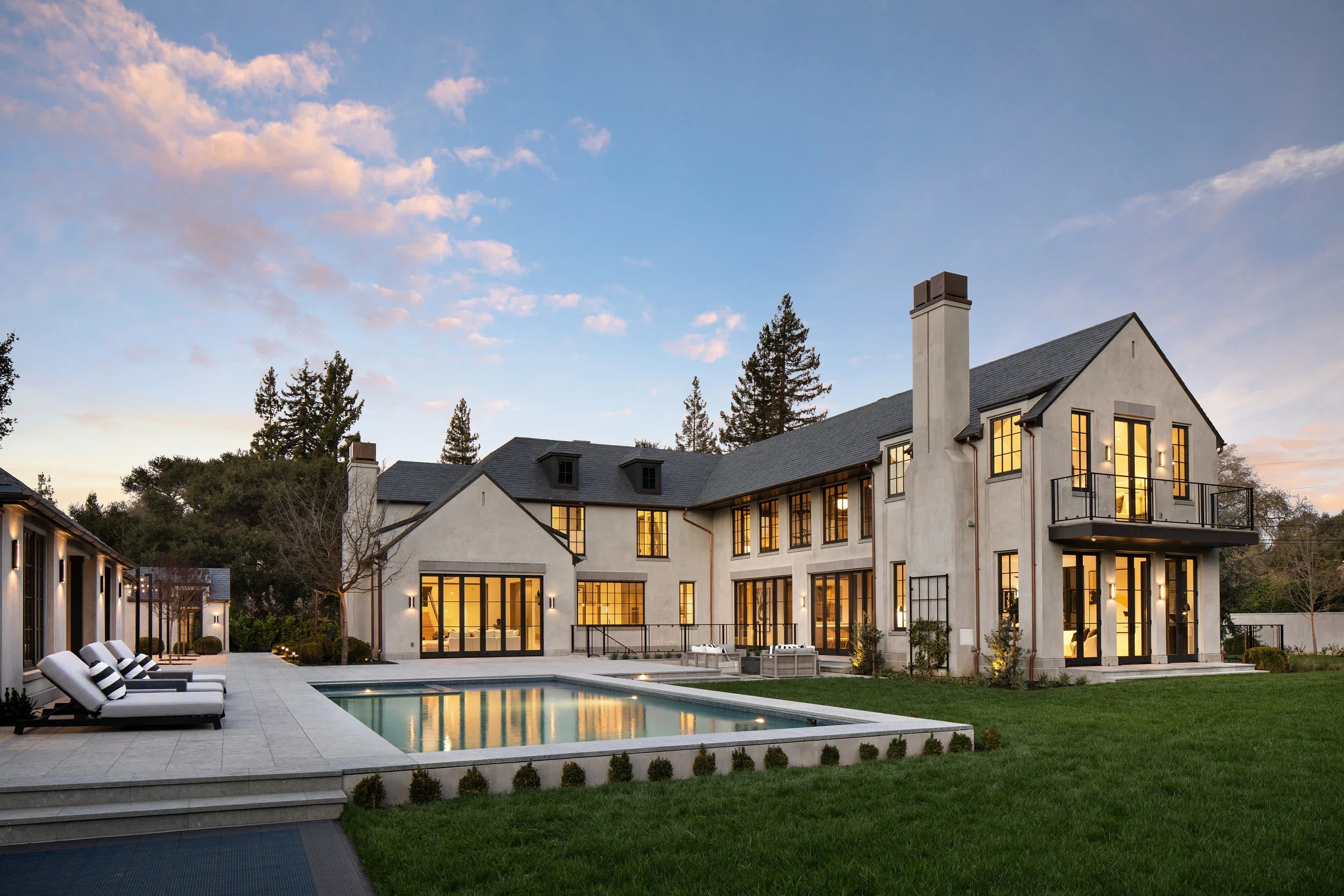 An exterior rear view of the residence at dusk, featuring a swimming pool, lounge chairs, a patio area, and mature perimeter trees.
