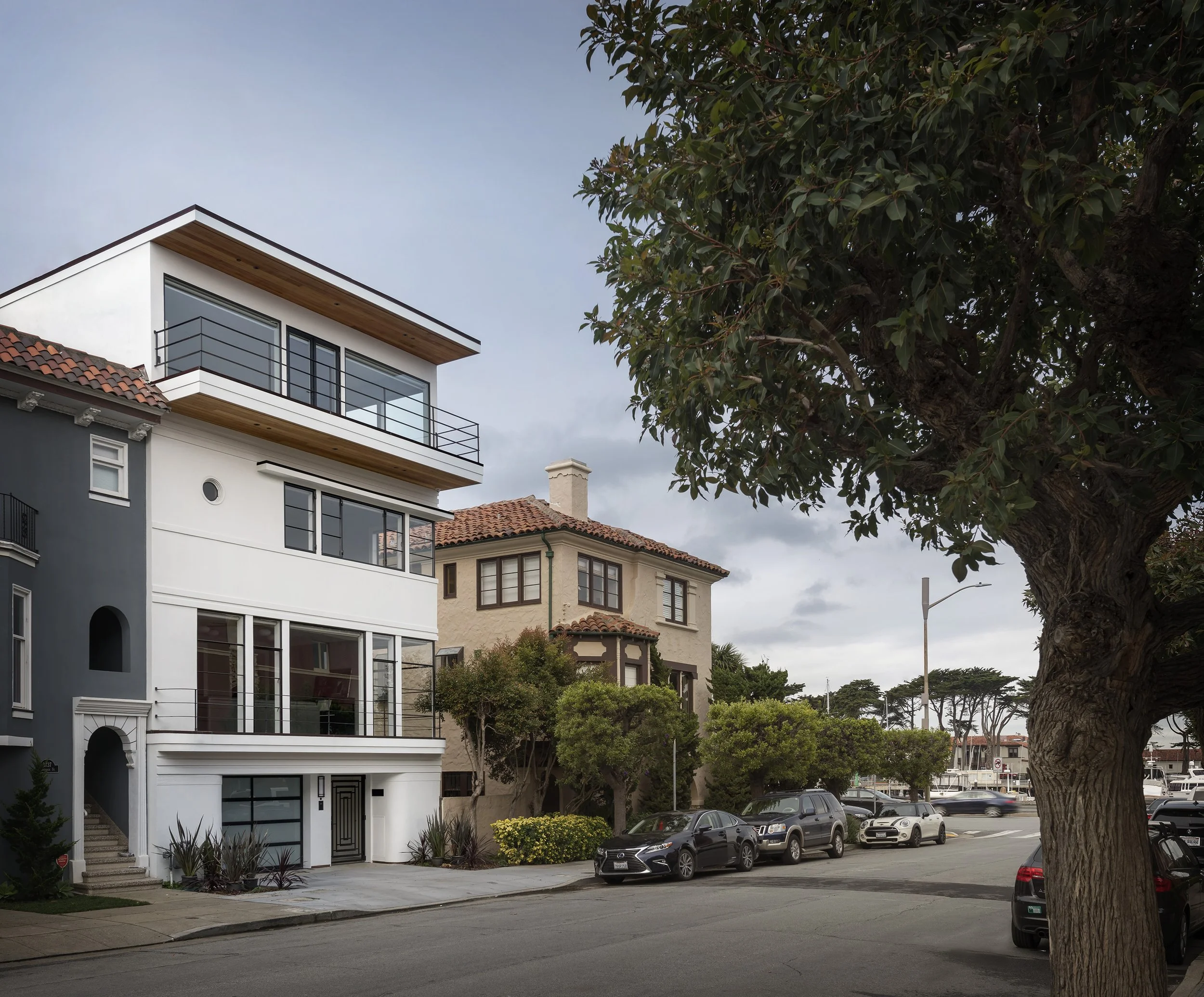 Exterior street-level view of a modern multi-story home with a flat roof, large windows, crisp white facade, a balcony with black railings, and mature street trees in front.