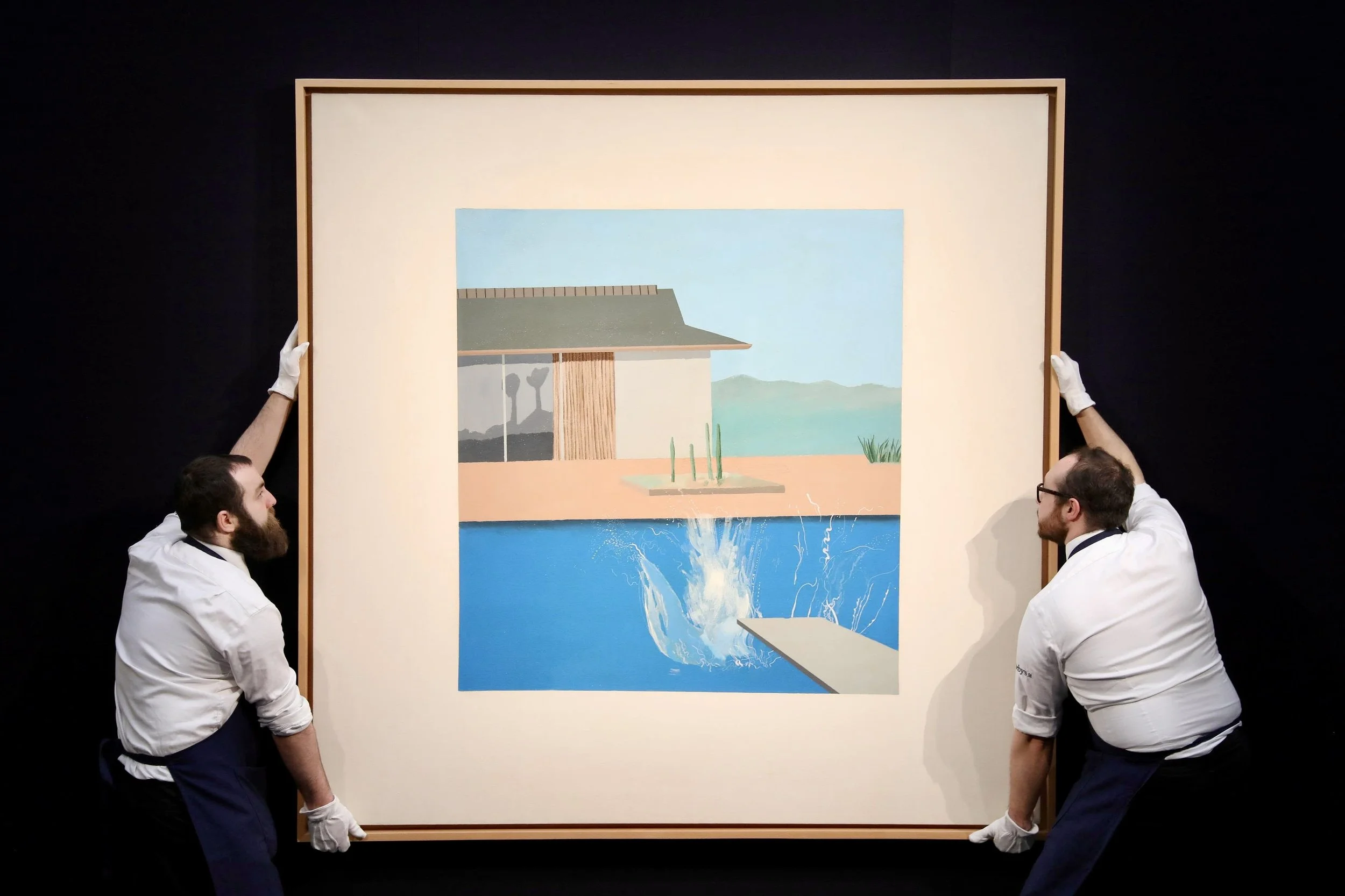 Two museum workers hold a large framed painting of a modernist house and swimming pool with a splash of water.