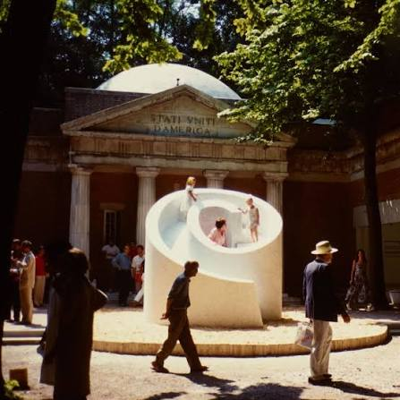 People gather around a large white sculptural slide installation in a historic courtyard shaded by trees.