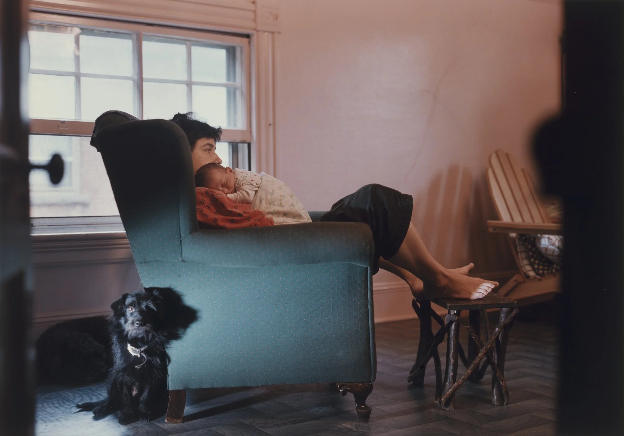A woman sits in a chair holding a baby, with a black dog sitting beside her in a warmly lit room.