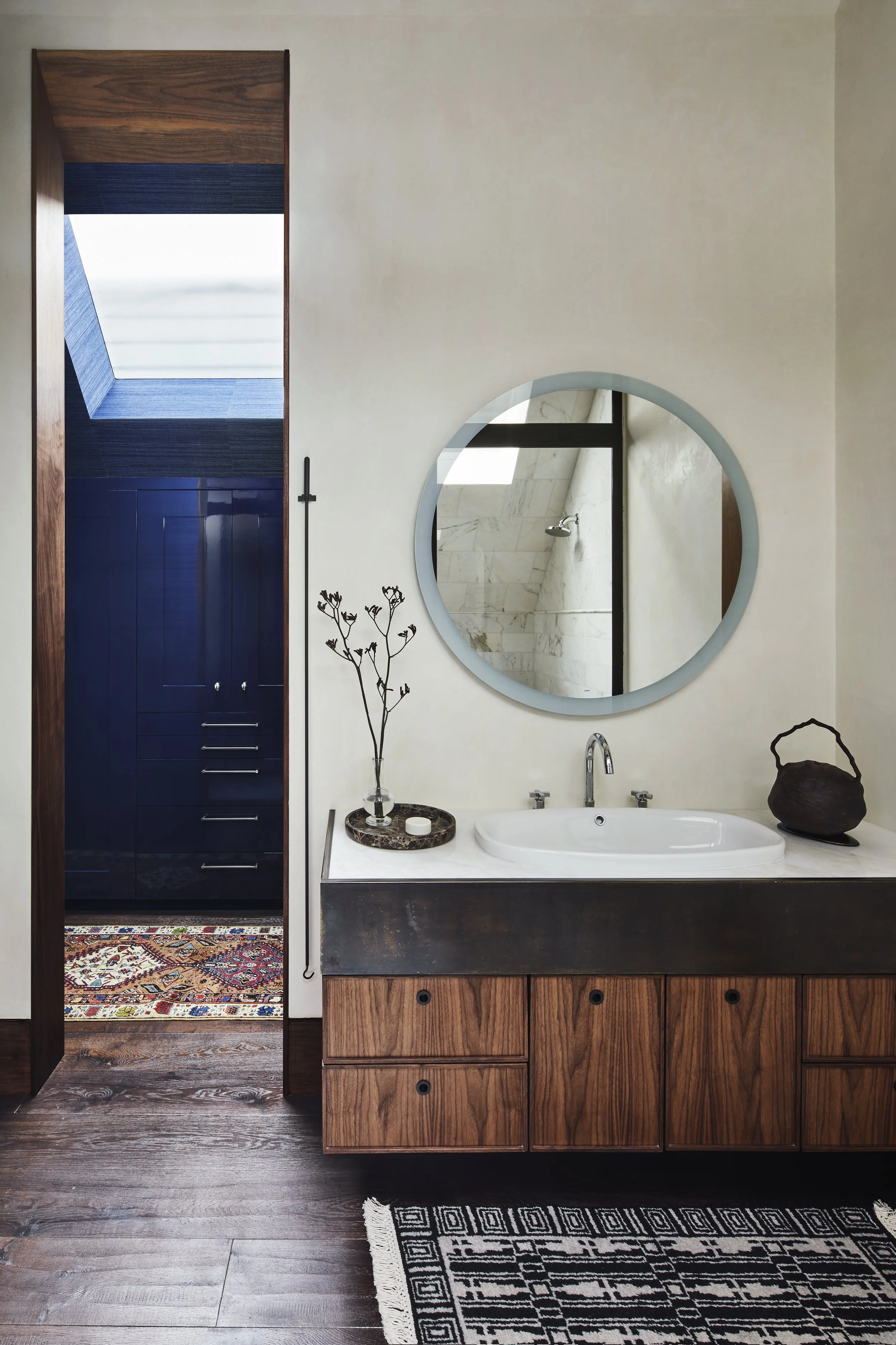 Bright, airy bathroom featuring a floating wooden vanity with a white countertop and basin sink, a round illuminated mirror, and an open door leading to a dark blue wall of floor-to-ceiling storage illuminated by a skylight.