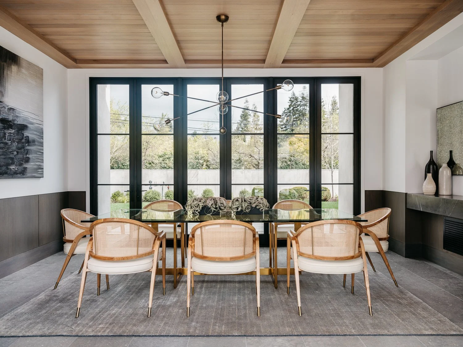 Interior view of the dining room furnished with a long dark table, light upholstered and caned chairs, a chandelier, and an open view of the surrounding landscaping.